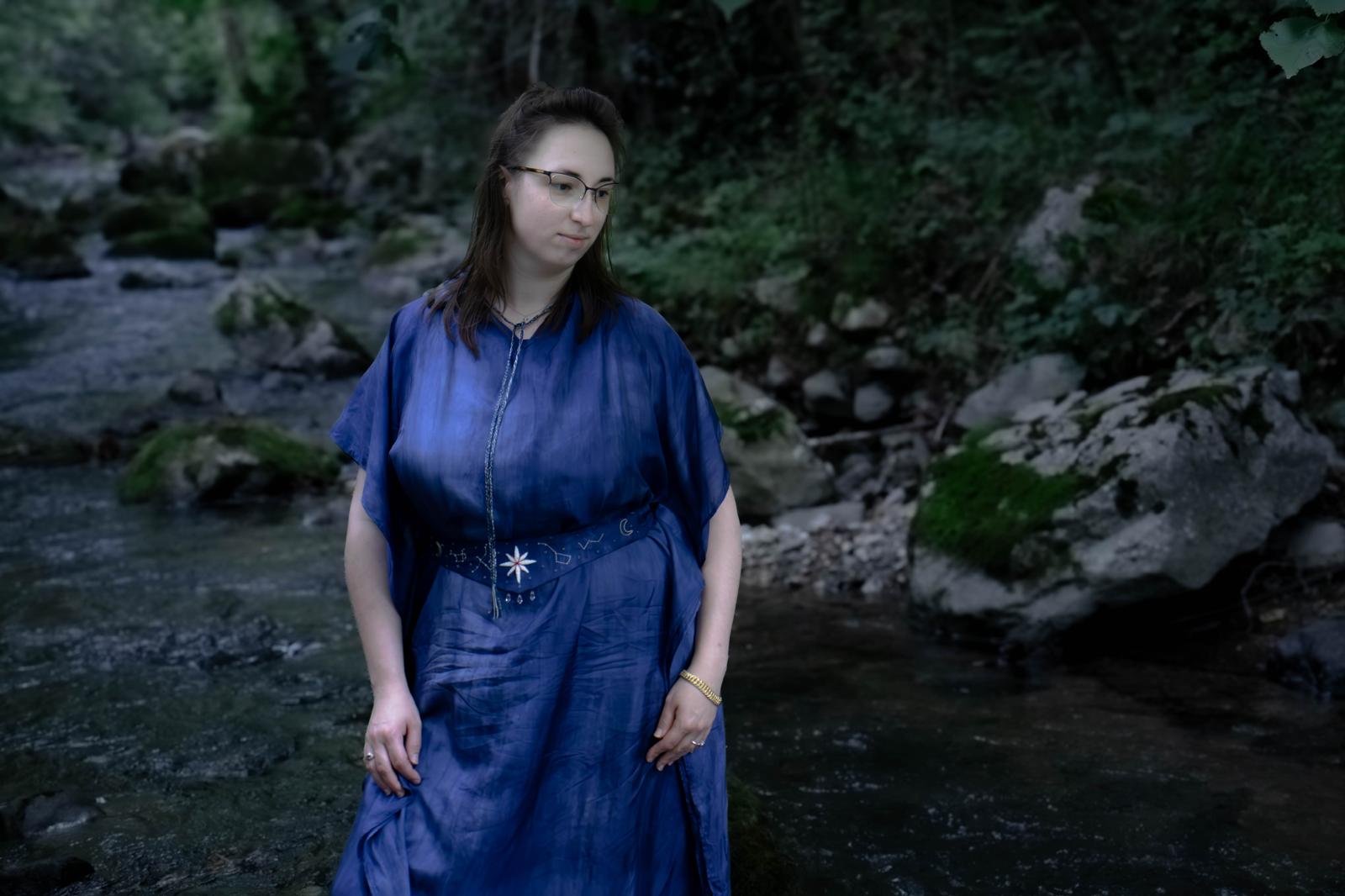 Une jeune femme portant une robe bleue avec des motifs d'étoiles et de lunes, debout dans une rivière entourée de rochers et de végétation dense, avec des lunettes et une montre dorée.