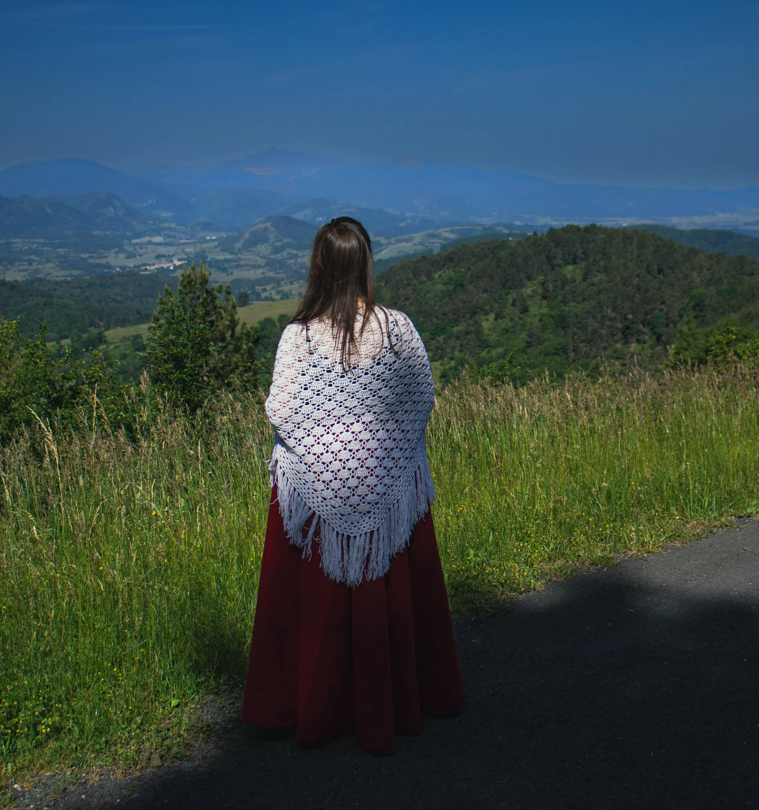 Femme vue de dos portant une robe longue rouge, avec un châle blanc crocheté, regardant un paysage de collines verdoyantes sous un ciel bleu avec montagnes à l'arrière-plan.