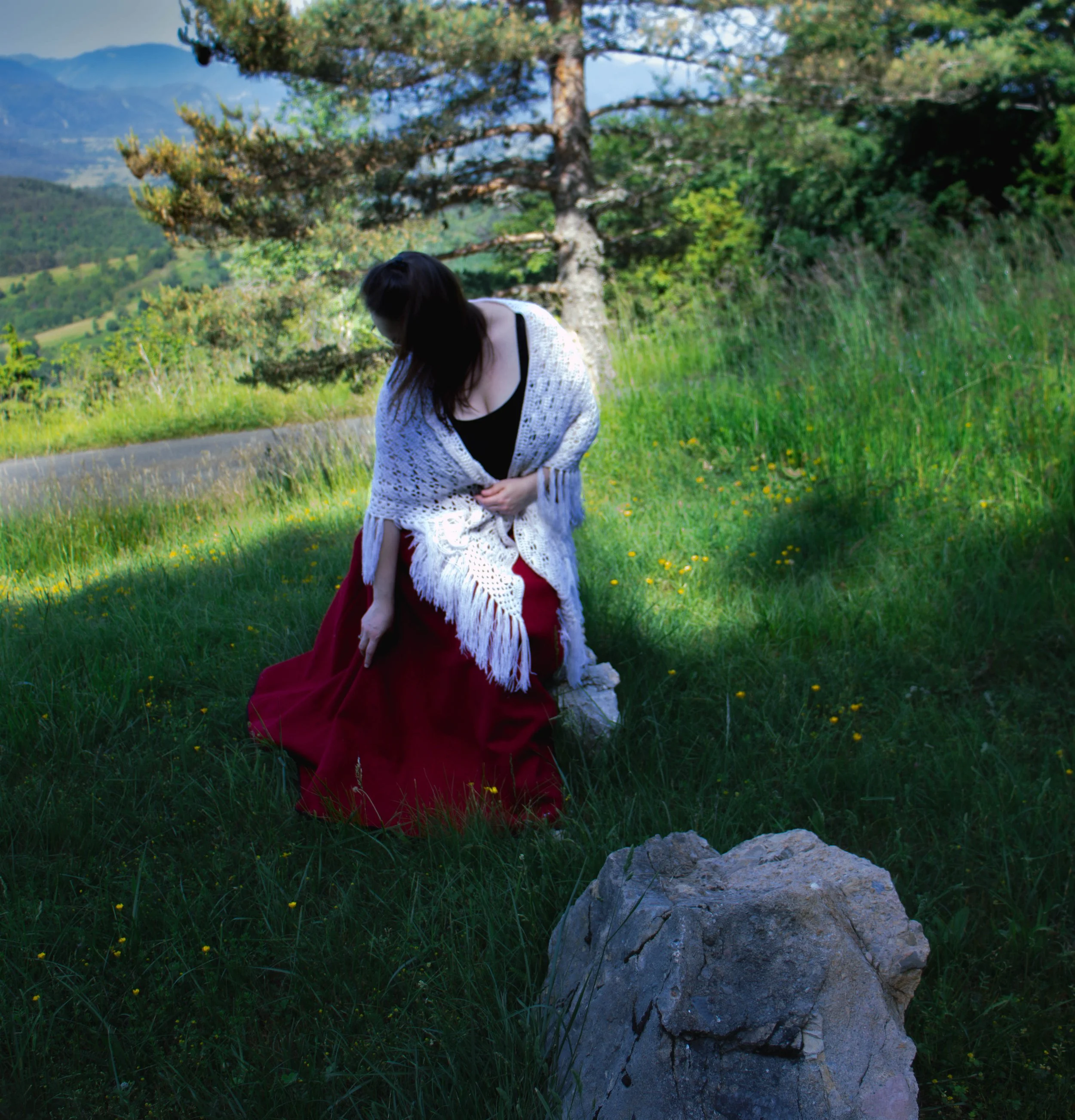 Une femme vêtue d'une robe rouge et portant une couverture blanche, debout dans un champ de fleurs et d'herbes vertes, avec un arbre à proximité et des montagnes en arrière-plan sous un ciel partiellement nuageux.