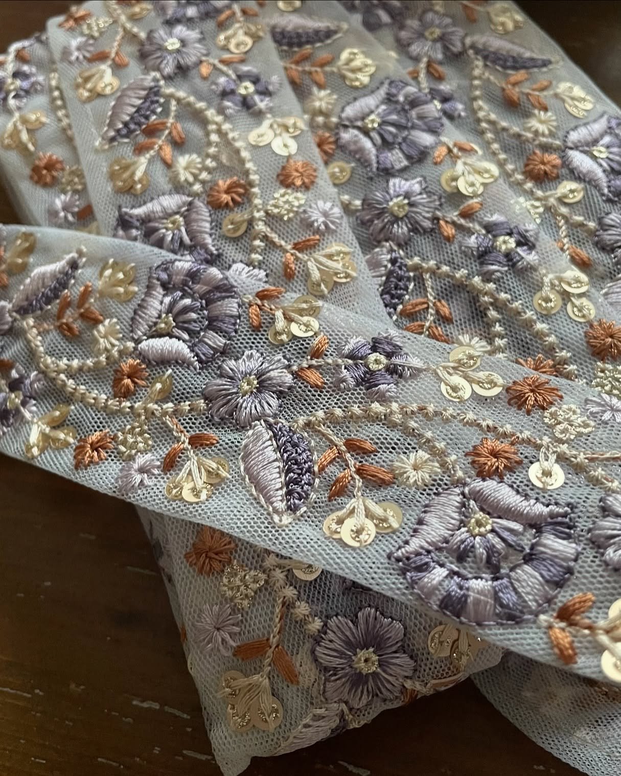 Tissu brodé avec des fleurs violettes, dorées et brunes, avec des sequins dorés, sur une grille fine. Style élégant et travaillé.