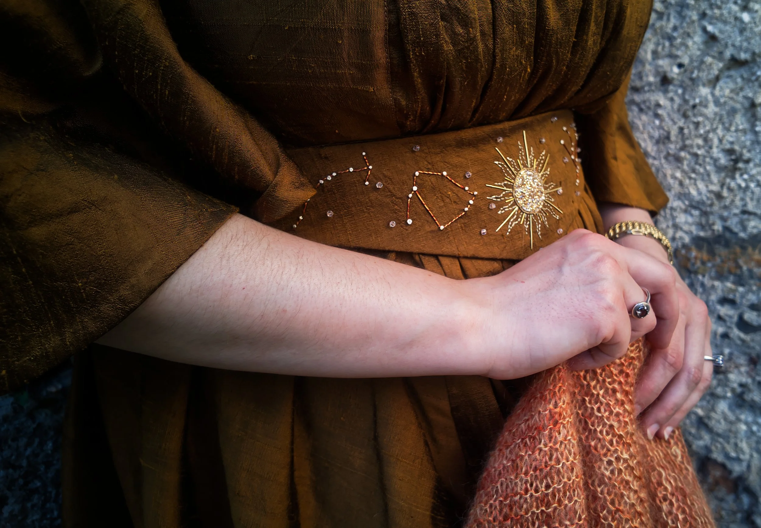 Manchette en tissu marron avec broderies de constellations et un soleil en or, mains croisées, bracelet en or, arrière-plan de roche grise.