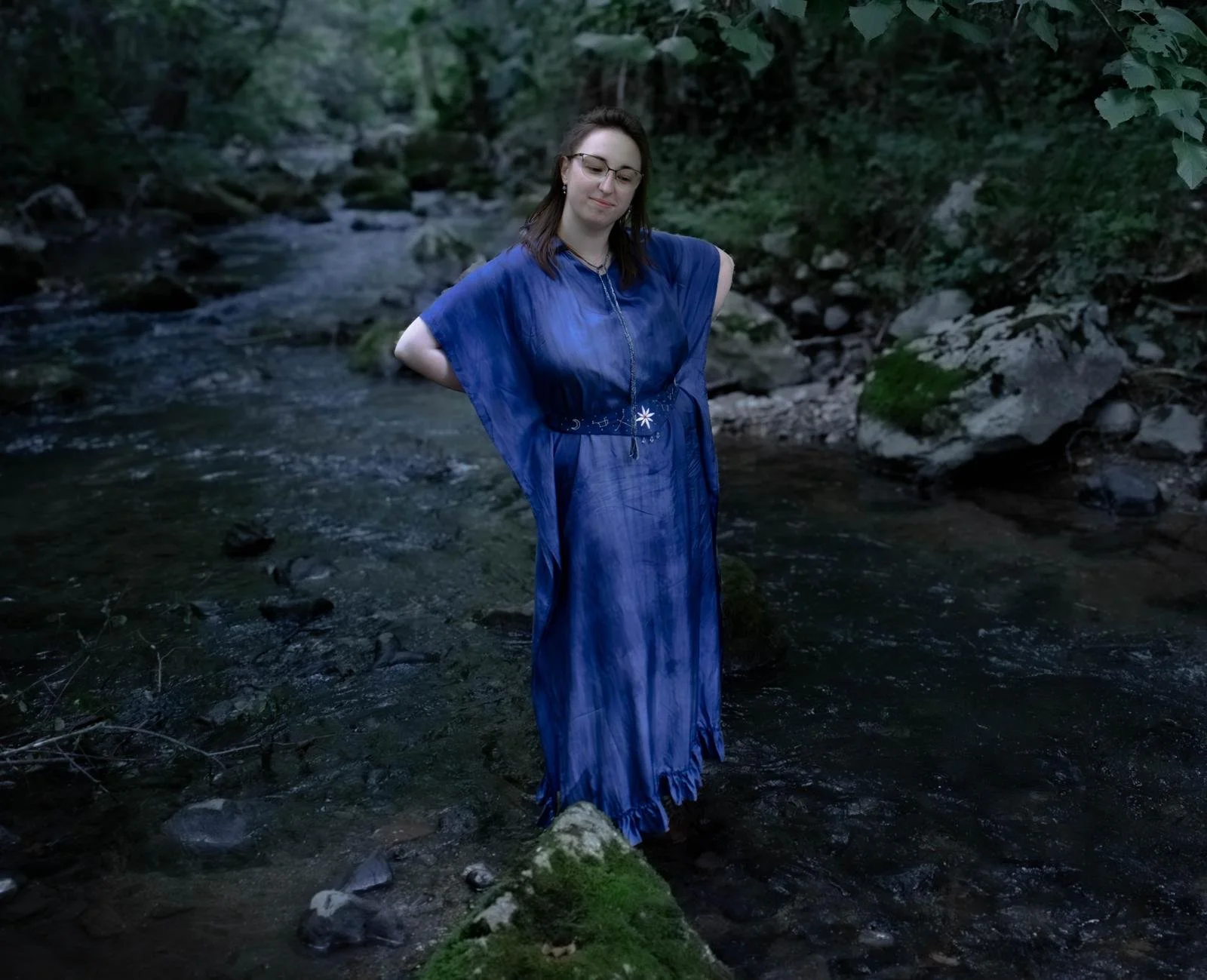Une femme en robe bleue se tenant dans une rivière entourée de végétation dense.