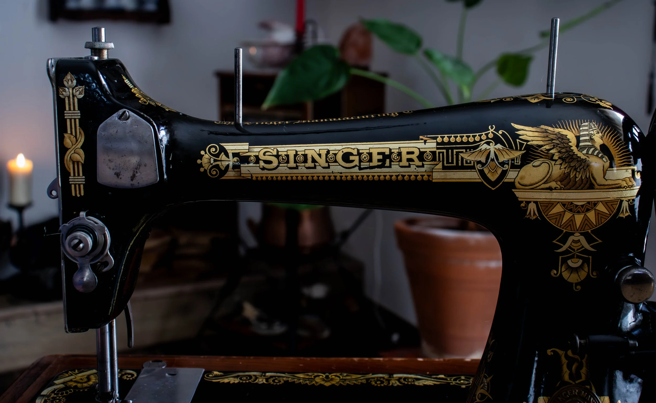 Machine à coudre noire avec des décorations gold sur le corps, portant le nom 'SINGER', dans un intérieur avec des plantes vertes et une bougie allumée en arrière-plan.