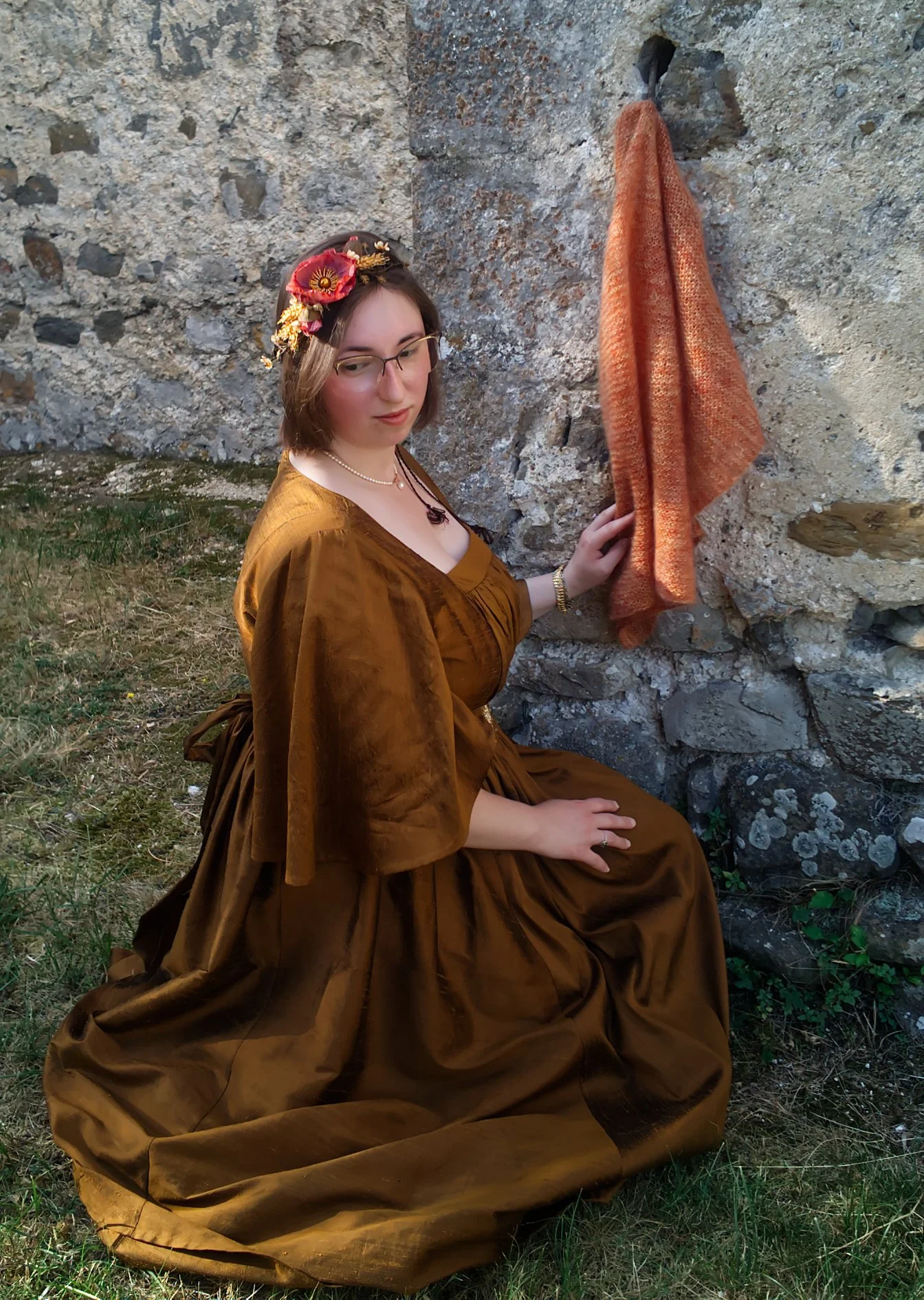 Une jeune femme habillée d'une robe marron en velours, assise sur l'herbe près d'une pierre, portant un bandeau de fleurs dans les cheveux. Elle touche doucement une écharpe orange suspendue à la roche.