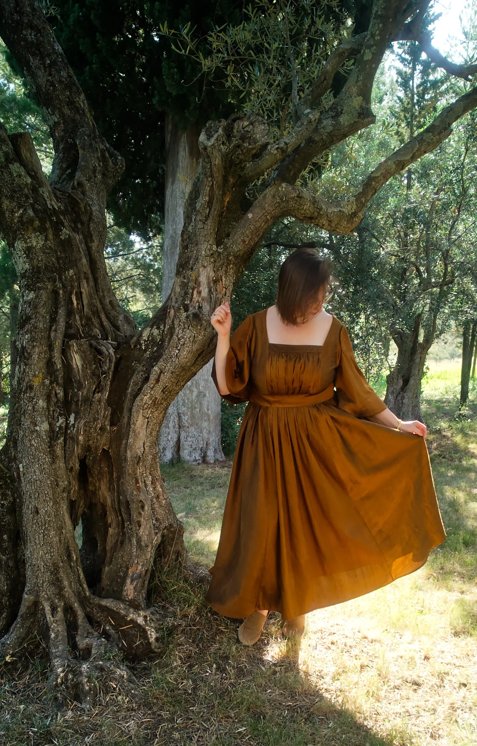 Une femme vêtue d'une robe marron, debout sous un arbre lors d'une journée ensoleillée dans un environnement naturel.