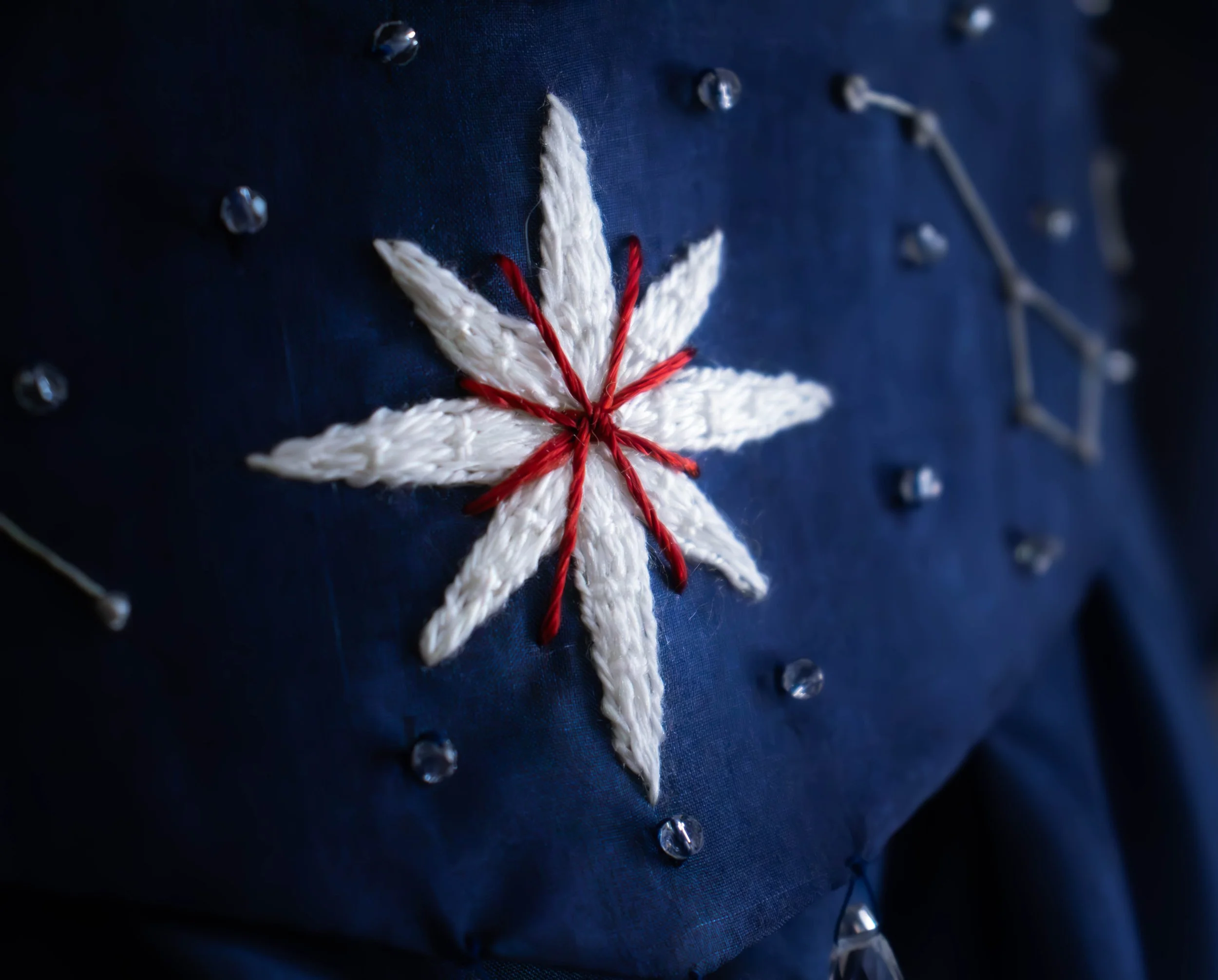 Broderie brodée en forme de fleur avec des fils rouges et blancs sur un tissu bleu foncé, avec des perles transparantes autour.