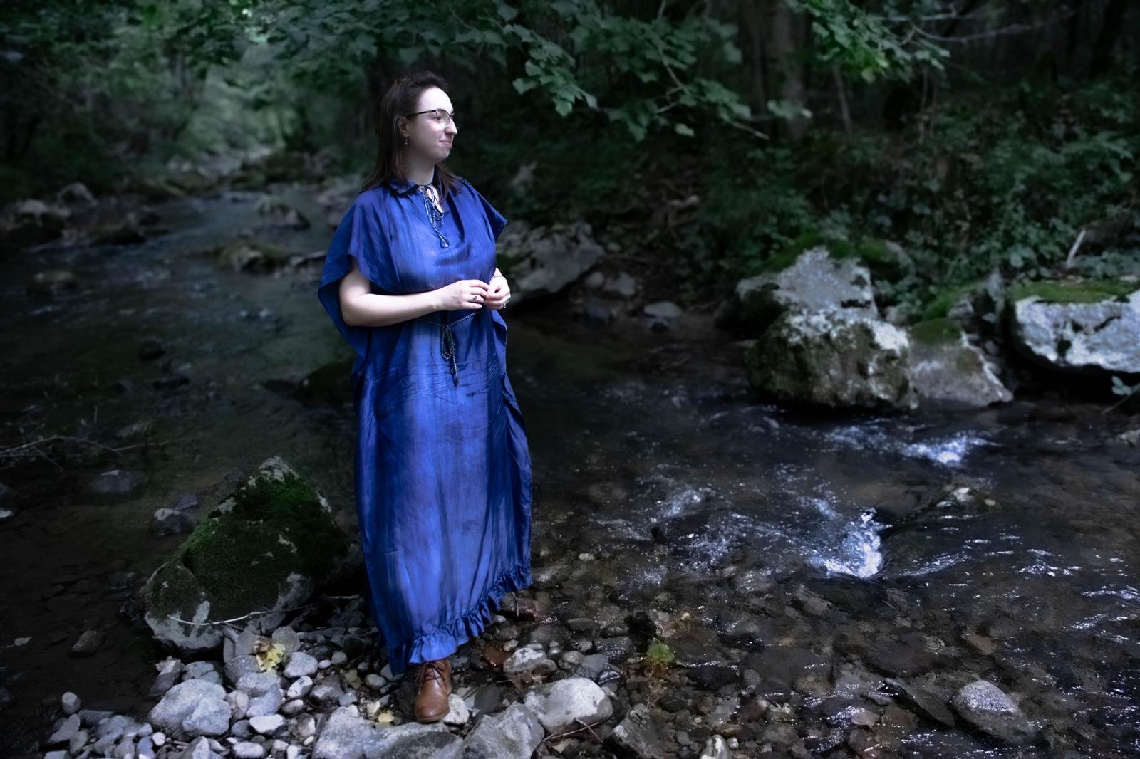 Une femme debout dans une rivière entourée d'arbres, portant une robe longue bleue et des lunettes. Elle regarde vers la gauche, paisible, dans un environnement naturel.