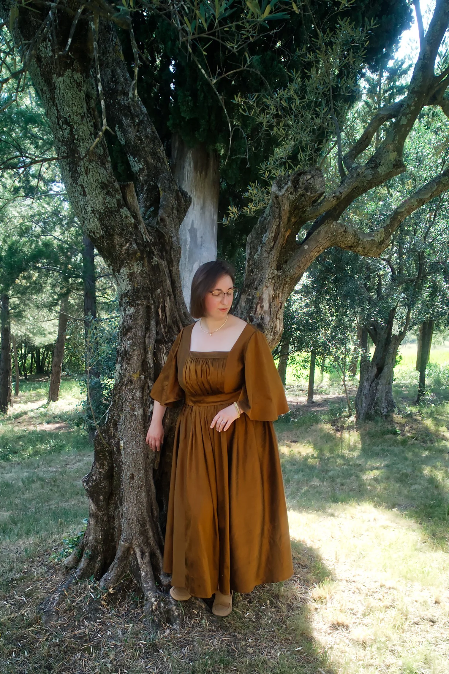 Une femme portant une robe marron olive, avec un collier blanc, se tient devant un arbre dans un jardin ensoleillé, entourée d'arbres verts et d'herbe.