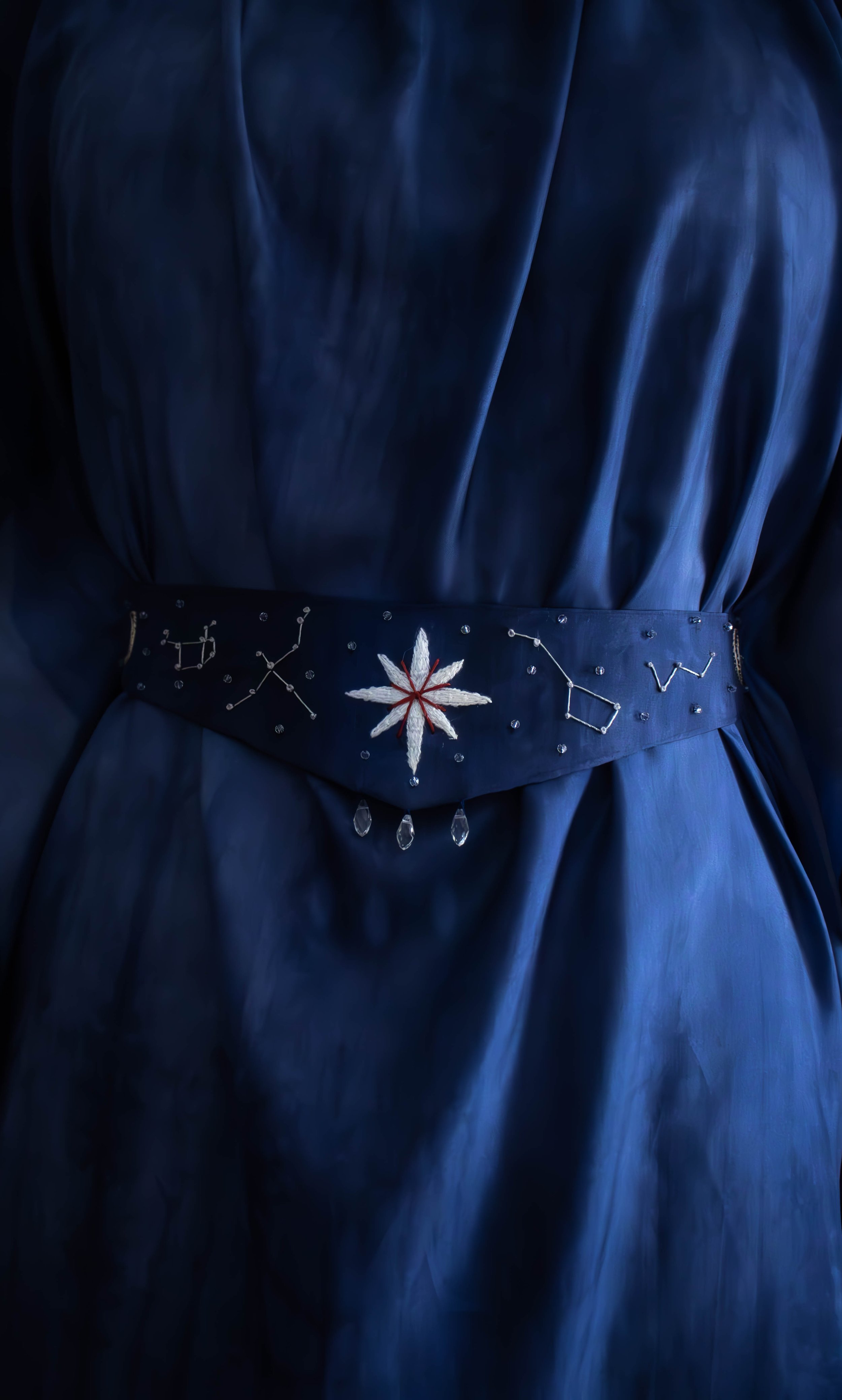 Ceinture ornée d'une étoile blanche en relief au centre, de petites perles et de constellations en fil métallique, portée sur une robe en tissu bleu foncé. 