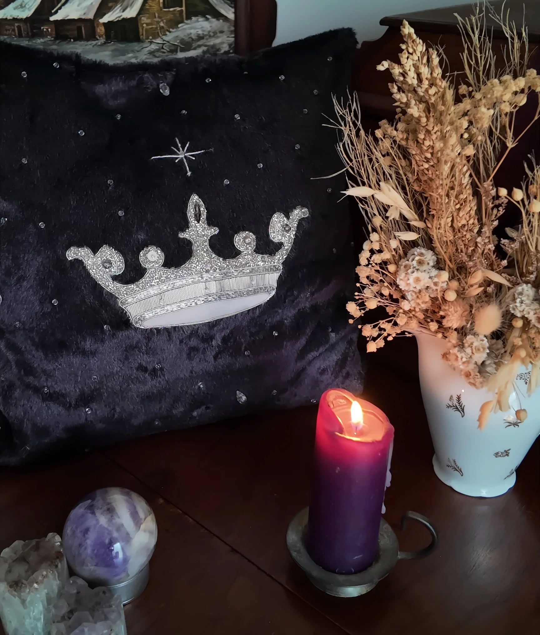 Une pièce décorée avec des objets de fête, une bougie violette allumée, un bouquet de fleurs séchées dans un vase blanc, et une nappe noire avec des détails brillants.