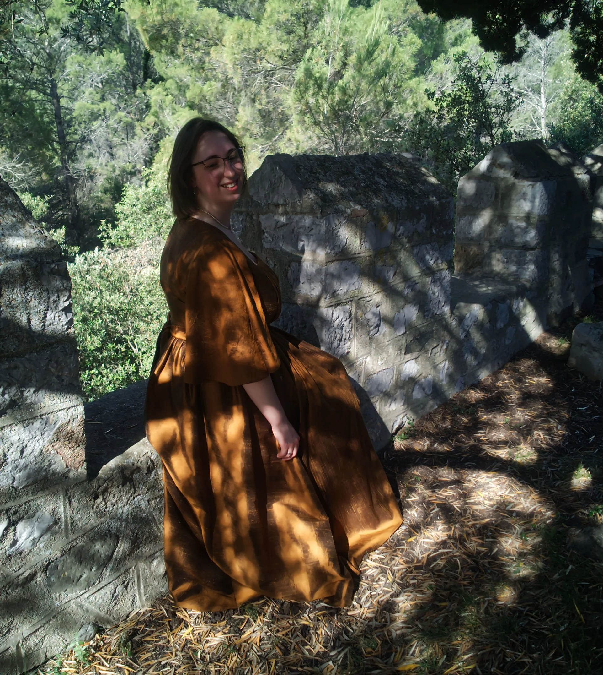 Une femme portant une robe en satin marron foncé se trouve près de rochers et de verdure dans une forêt ensoleillée.