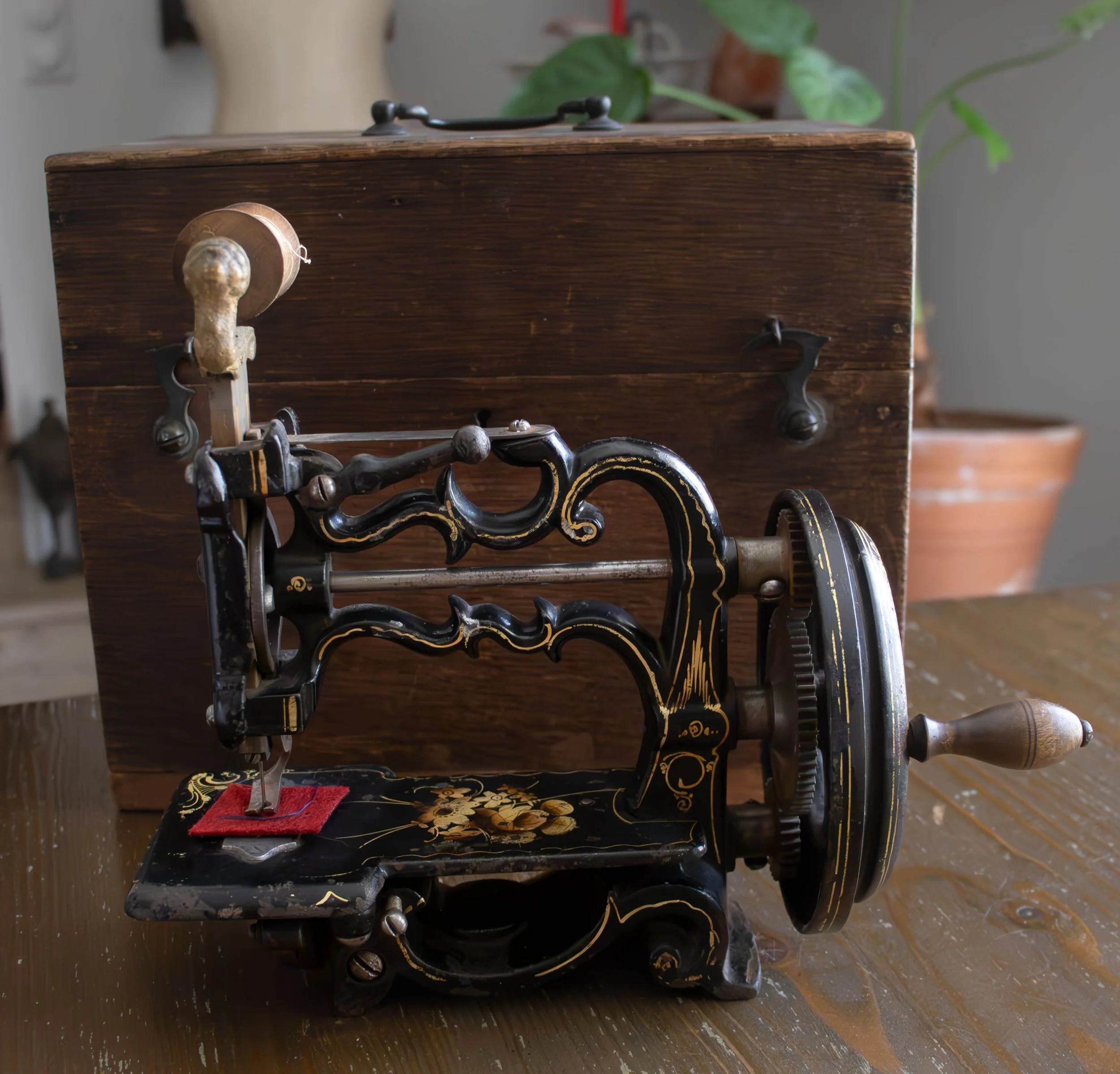 Une machine à coudre ancienne noire avec des accents dorés, posée sur une surface en bois, avec une petite pièce rouge en tissu sous la nappe, et des plantes vertes en arrière-plan.