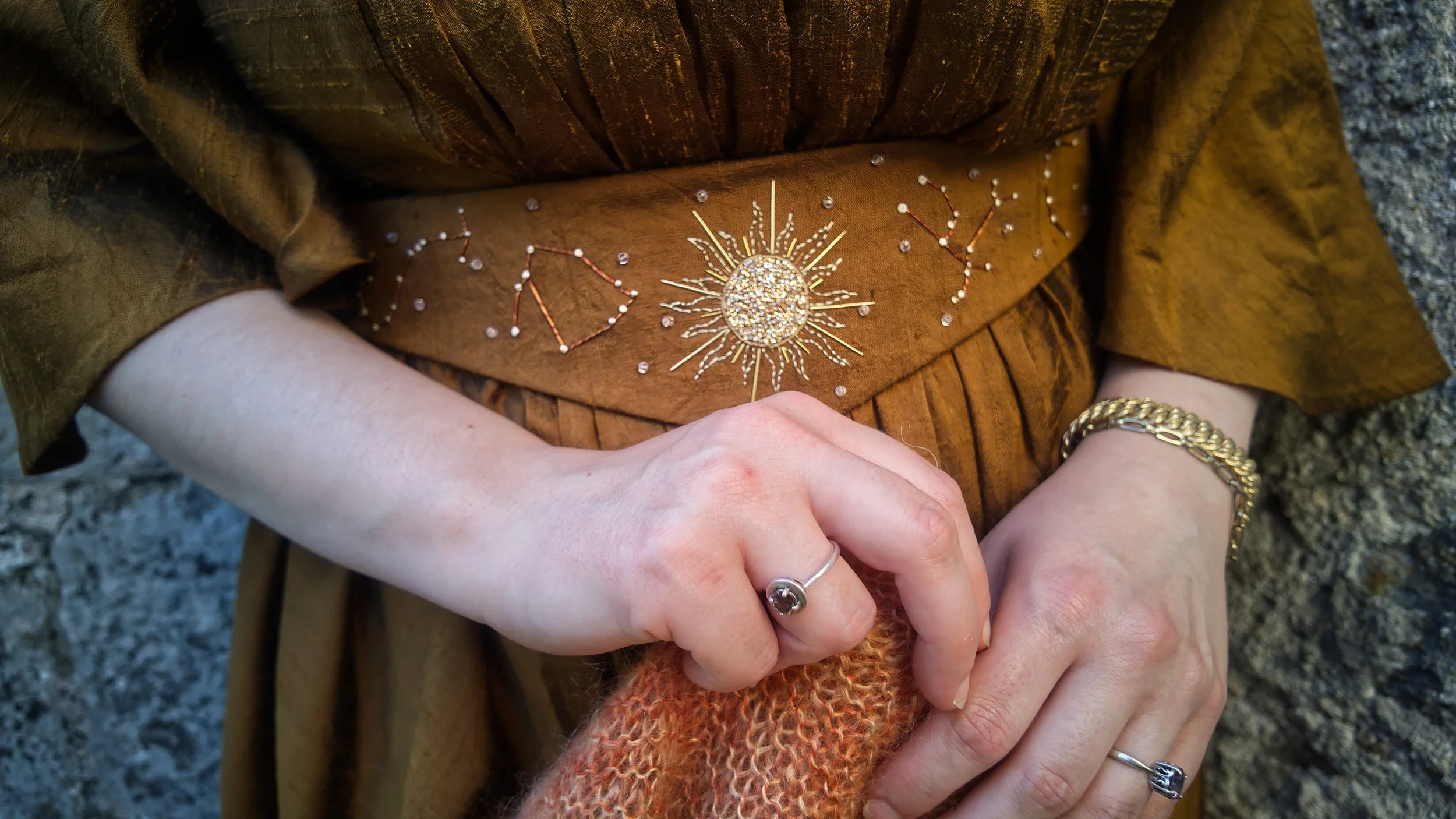 Une personne porte une veste en cuir marron avec des broderies de constellation et un soleil brillant, ses mains croisées portent des bijoux en or et en argent.