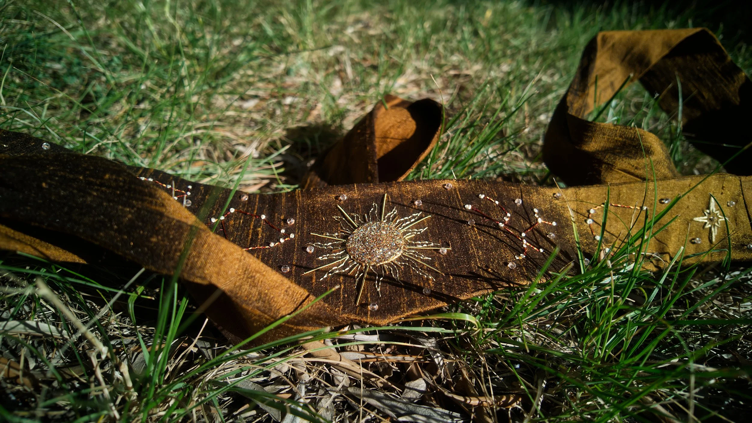 Une feuille morte avec un pendentif en forme de soleil brillant posé dessus, entourée d'herbes vertes et de petits éléments brillants.