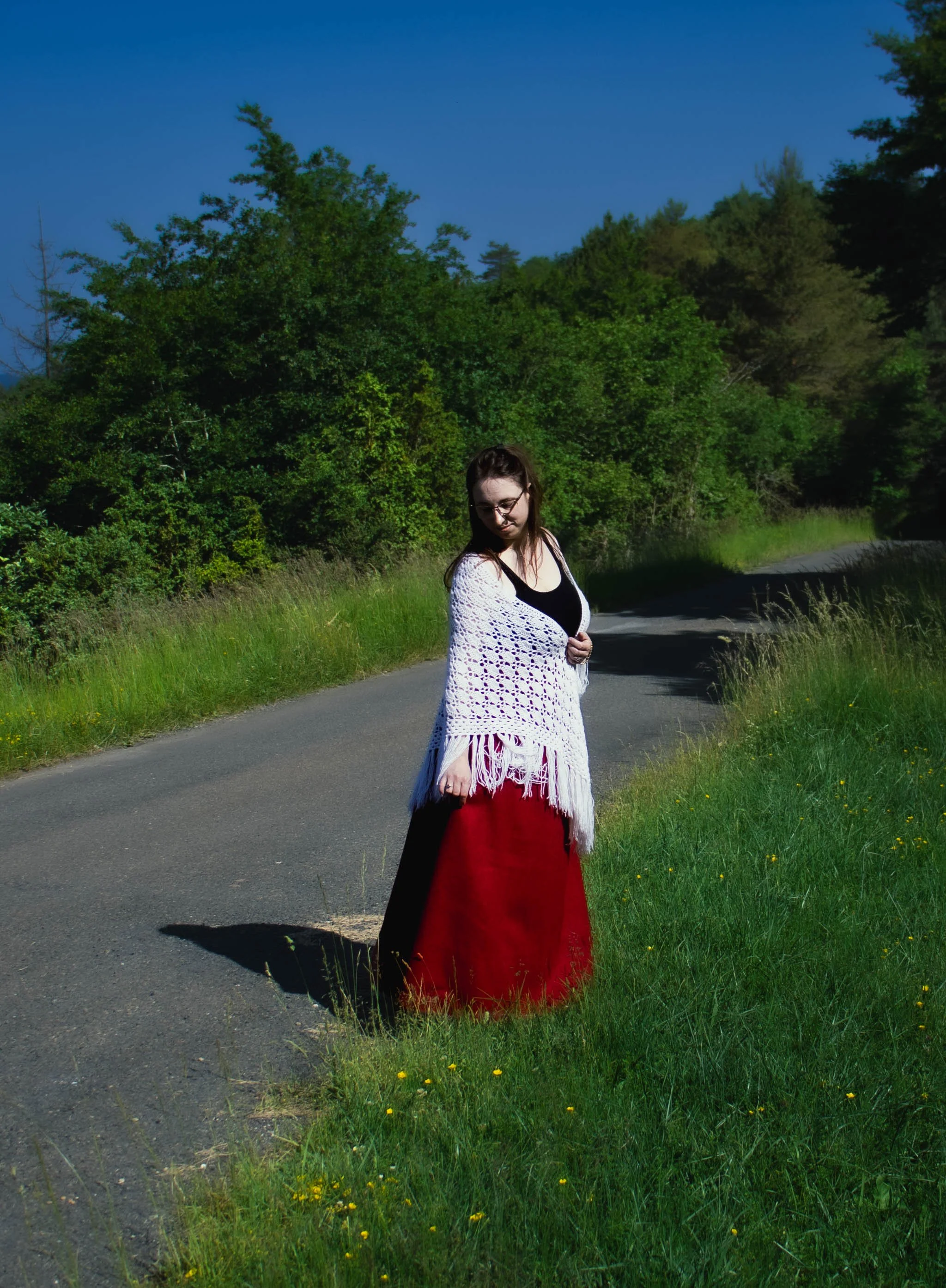 Une jeune femme portant une robe rouge, une blouse blanche et des lunettes, debout sur un chemin rural entouré d'herbes vertes et d'arbres, sous un ciel bleu clair.