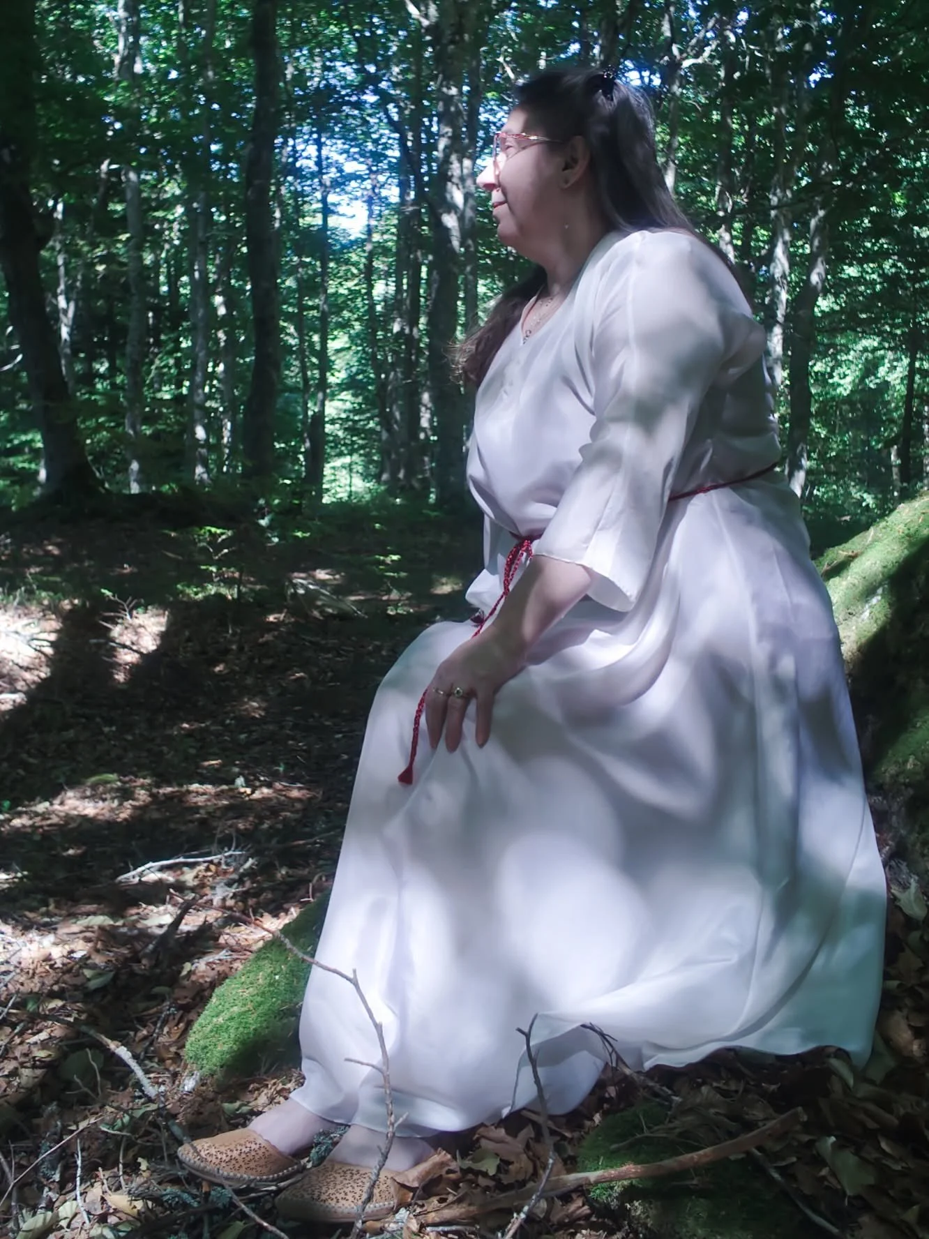 Femme assise sur une branche d'arbre dans une forêt, portant une robe blanche et des sandales à motifs, entourée de feuilles et de branches.