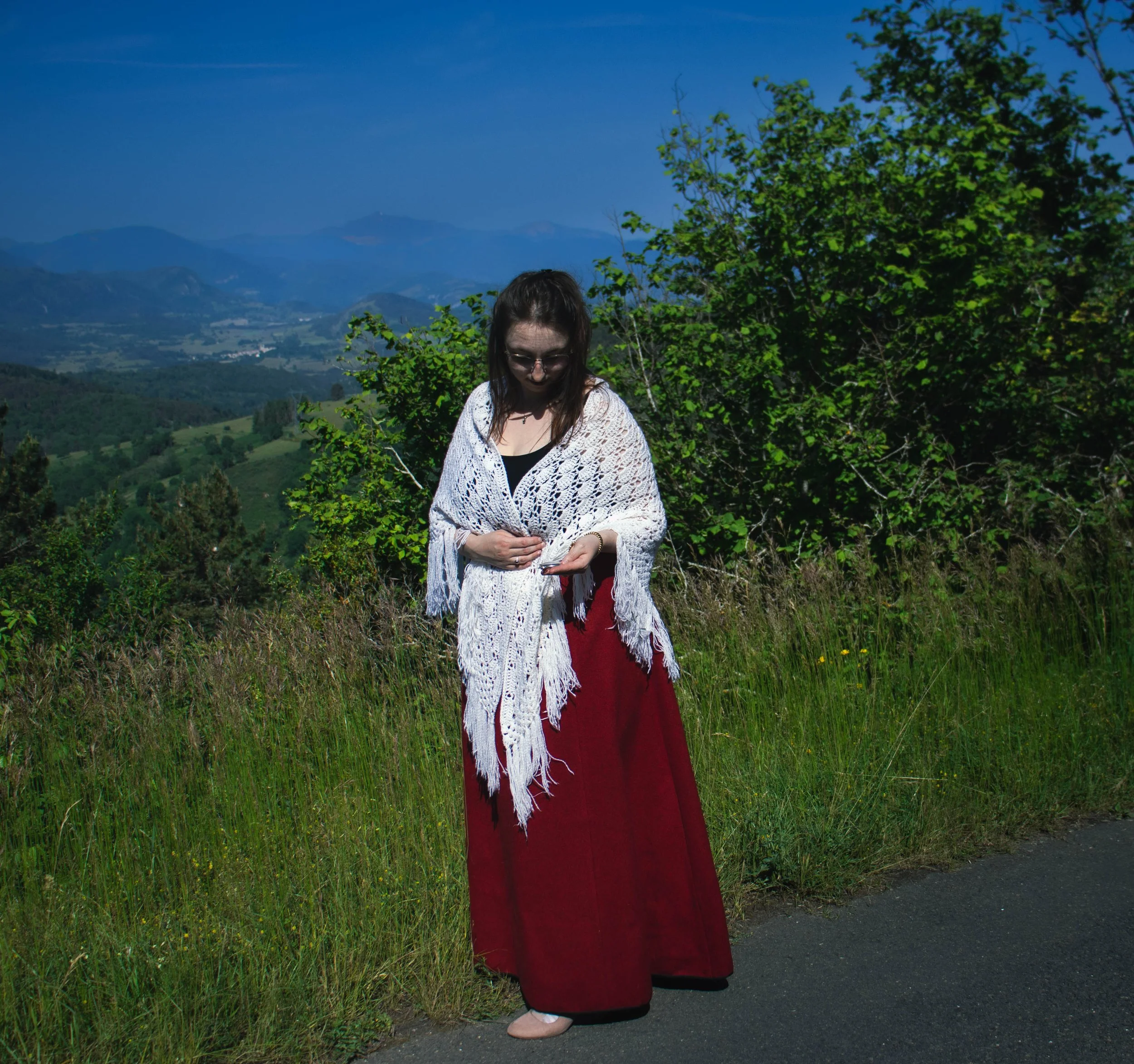 Une femme portant une robe rouge, une veste blanche en crochet et des chaussures blanches marche le long d'une route entourée de nature avec des montagnes en arrière-plan.