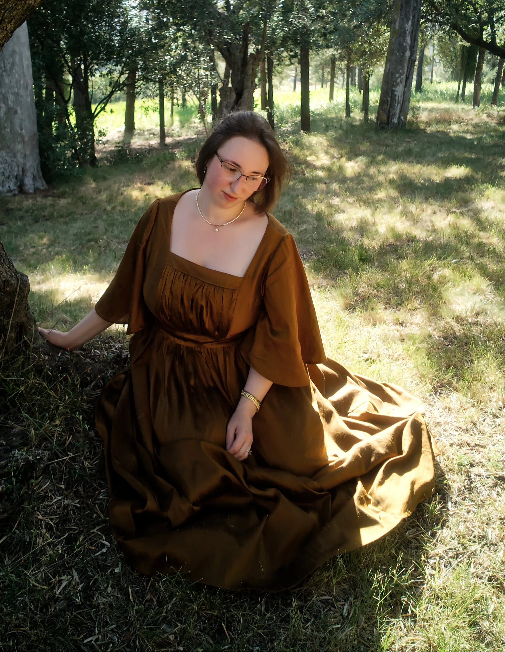 Jeune femme assise dans un parc ombragé en robes marron, portant des lunettes, une perle et un bracelet, entourée d'arbres et de verdure.