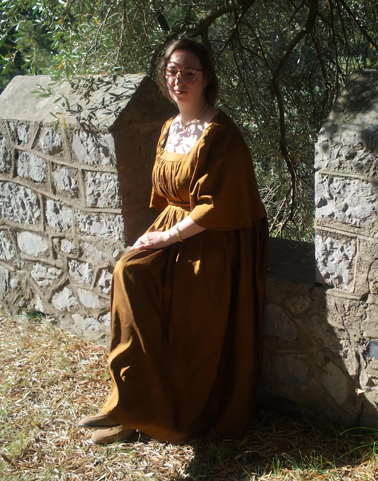 Femme assise près d'un mur en pierre, portant une robe marron vintage, sous un arbre avec de la lumière filtrée par les branches.
