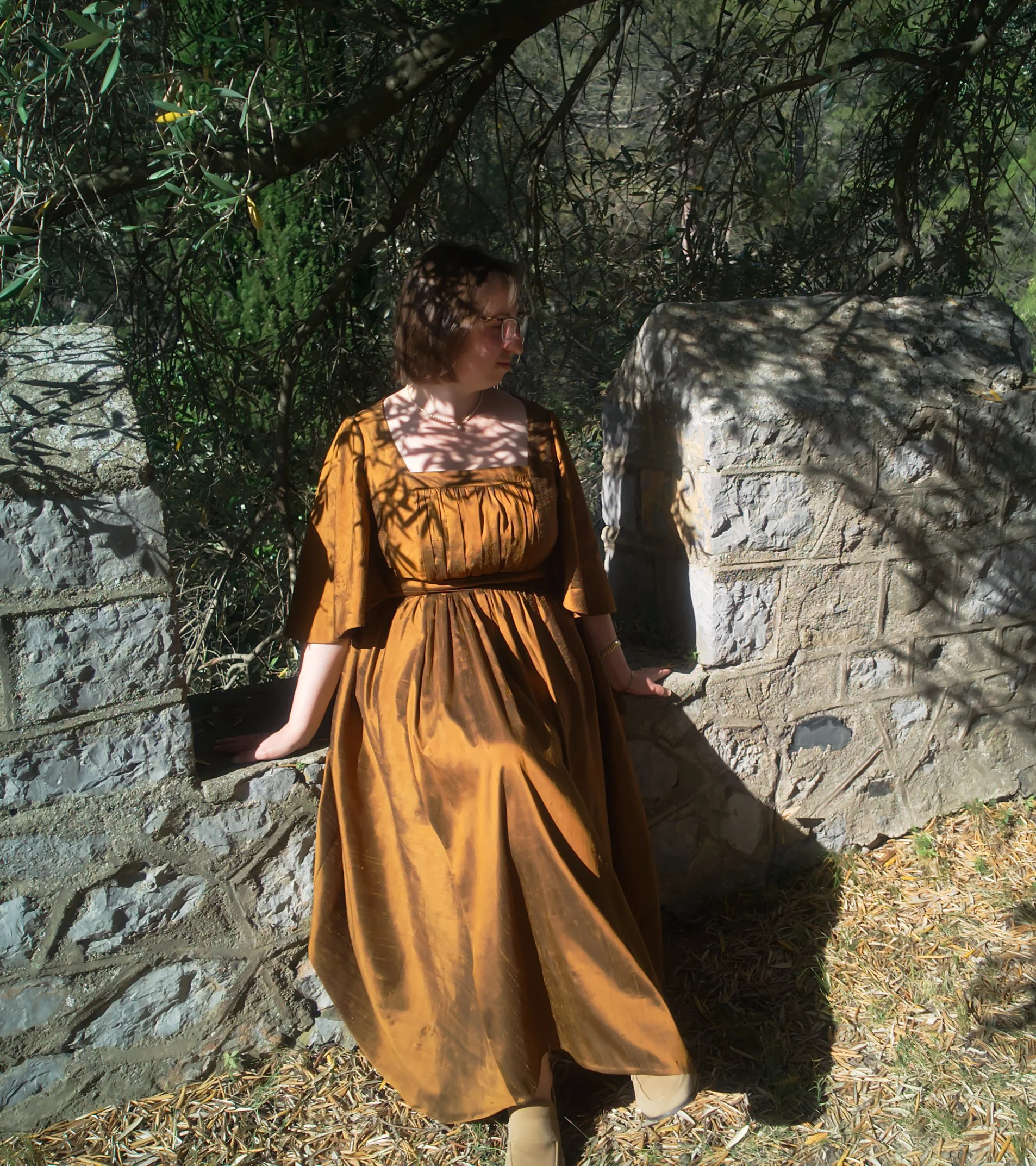 Une femme en robe marron clair se tient à côté d'un mur en pierre dans un environnement naturel avec des branches d'arbres et des feuilles. Elle porte des lunettes et regarde vers le bas.