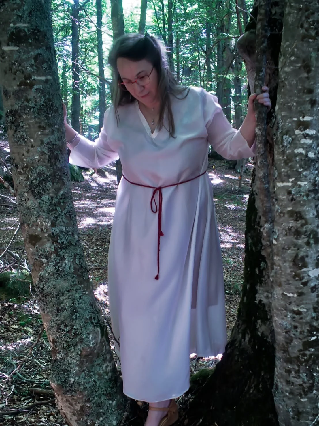 Une femme portant une robe blanche et des lunettes, debout dans une forêt, entre deux arbres, avec une expression concentrée.