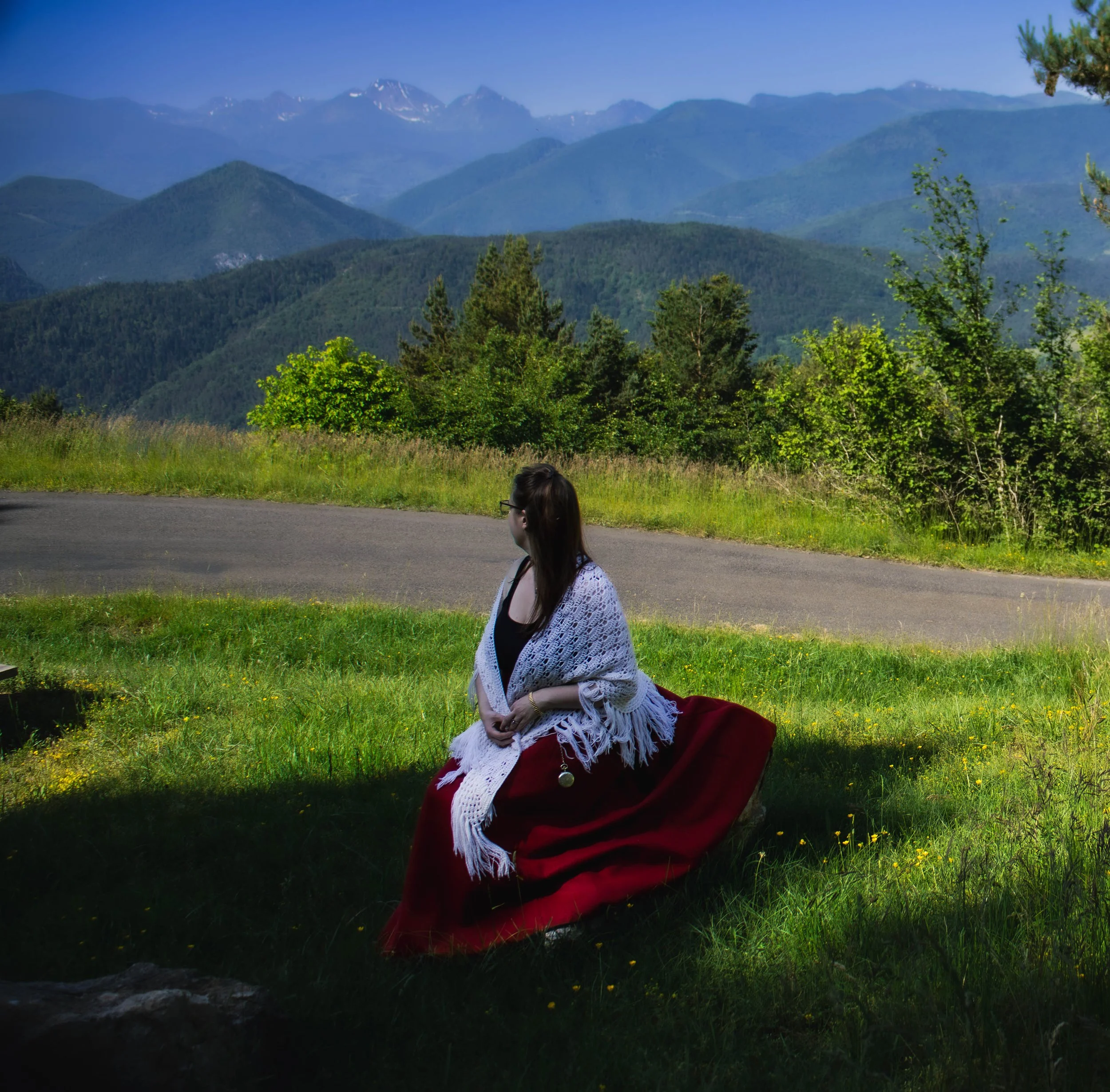 Une femme assise dans l'herbe verte près d'une route, avec des montagnes avec des sommets enneigés en arrière-plan, un ciel bleu clair et des arbres verts autour.