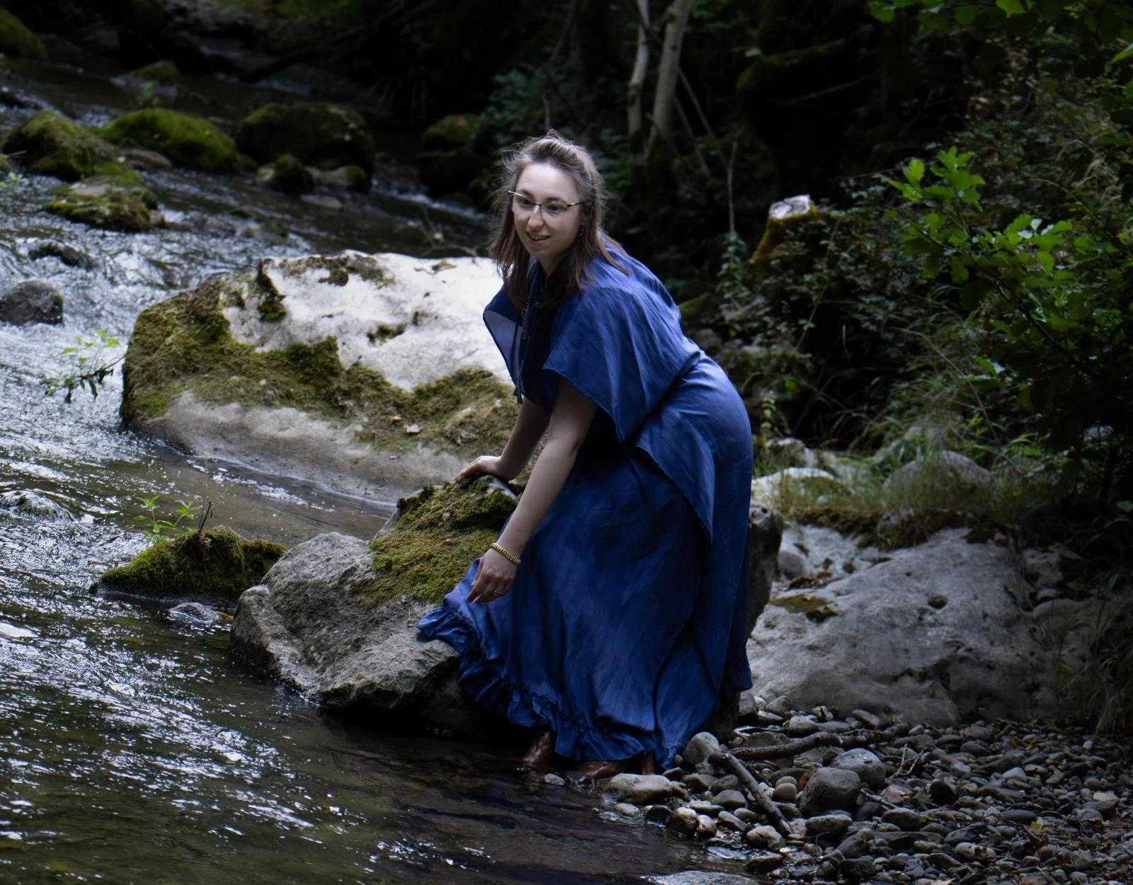 Une femme en robe bleue assise sur une pierre dans une rivière entourée de végétation dense.