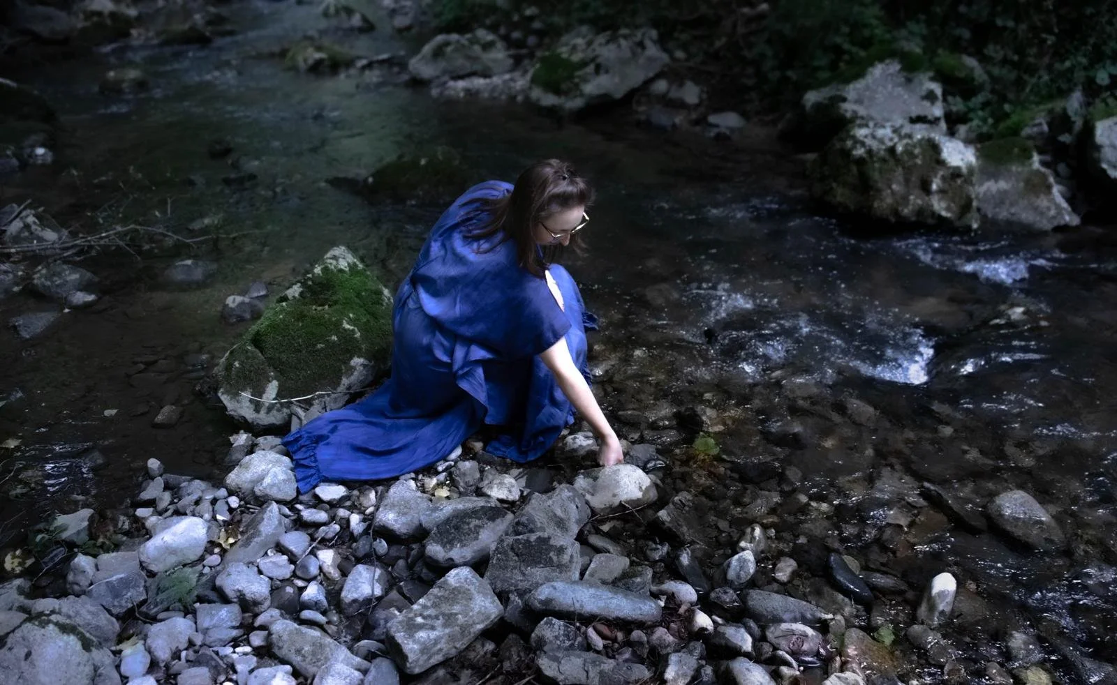 Une femme en robe bleue assise sur des rochers au bord d'une rivière en forêt.