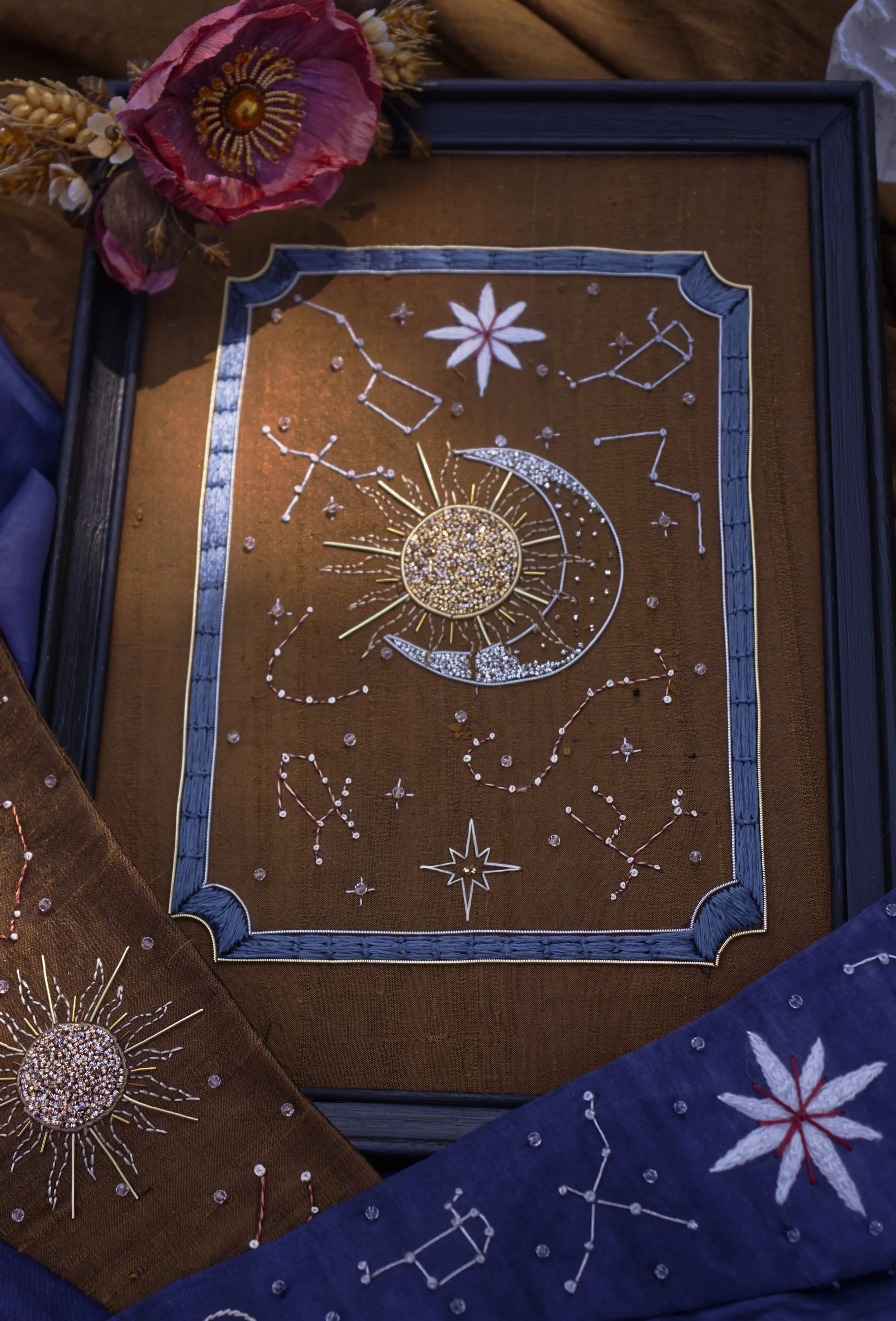 Tableau décoratif avec motifs de constellations et de symboles célestes, entouré de tissus en bleu et marron avec broderies de fleurs et de figures stellaires.