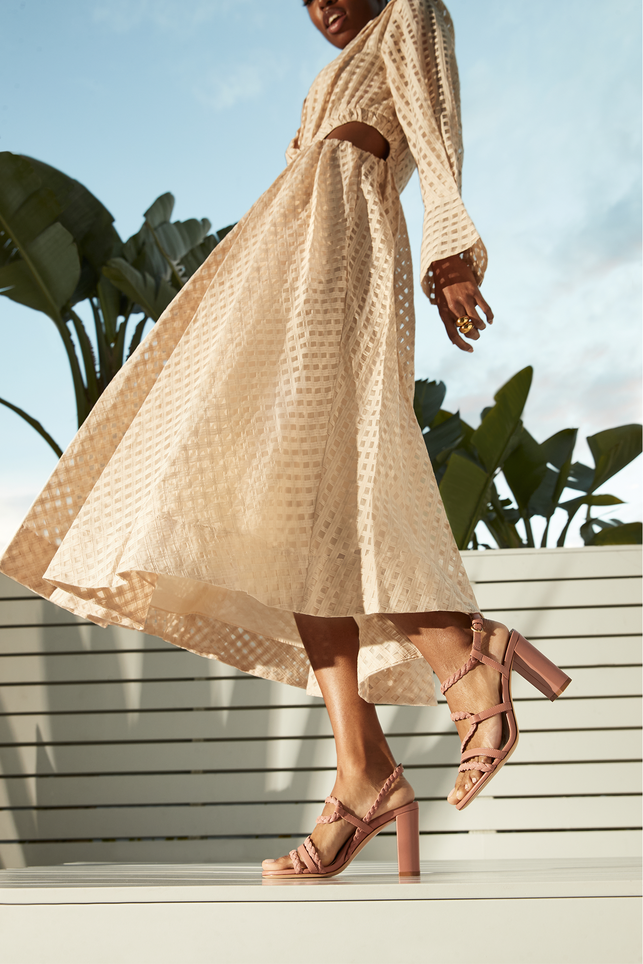 Close-up of a woman wearing a beige checkered dress with cut-outs, high-heeled pink sandals, and rings, standing outdoors against a blue sky with clouds and green plants.