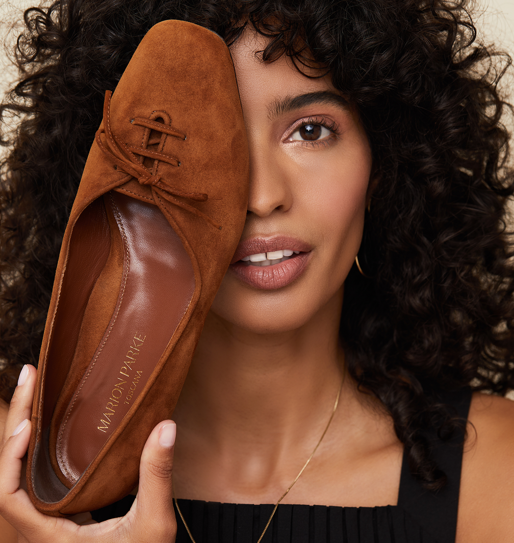 A woman with curly hair holding a brown shoe over her face, covering one eye, while smiling.