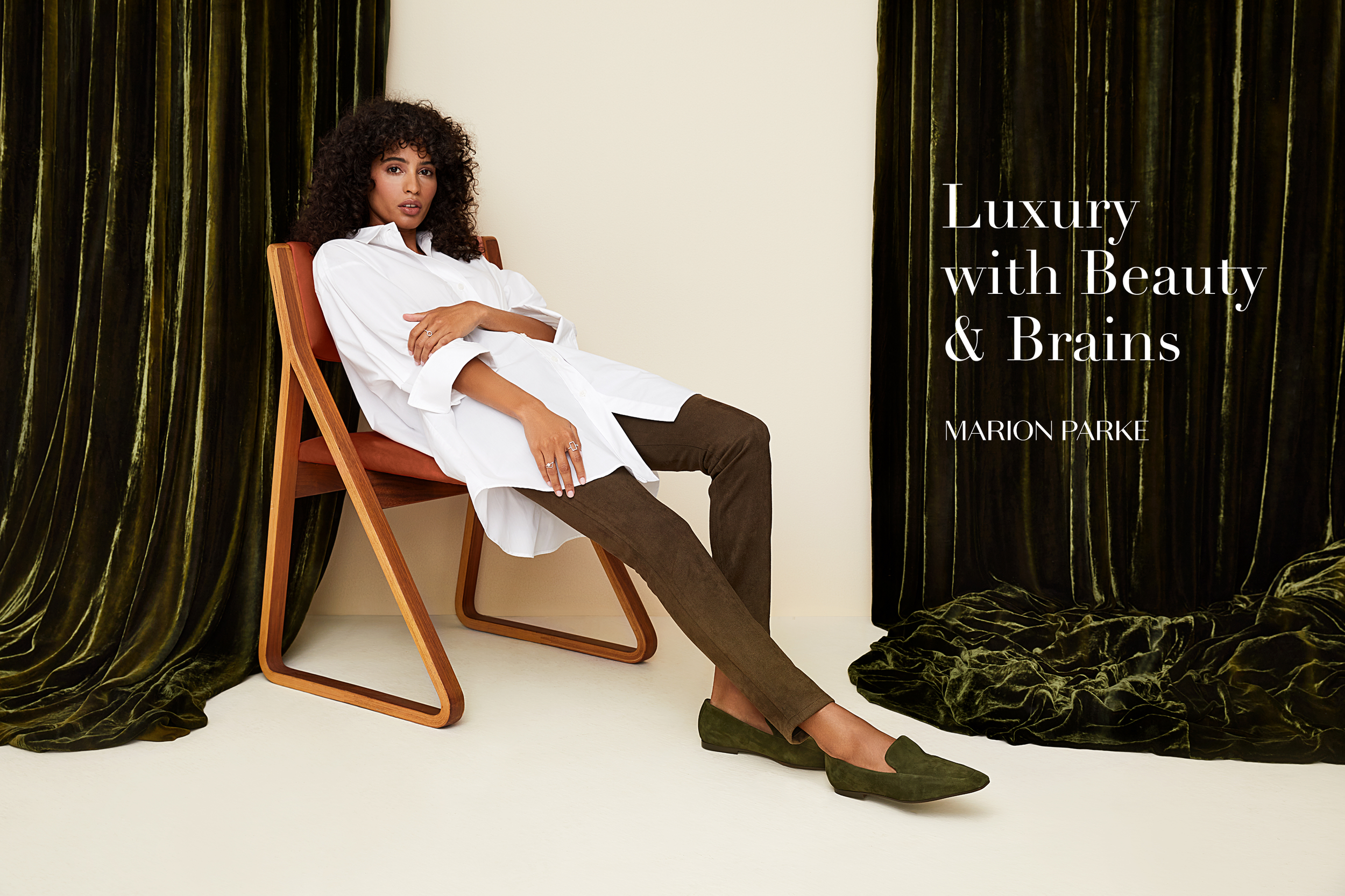 A woman with curly hair sitting on a wooden chair with a red cushion, wearing a white shirt and brown pants, in front of dark green velvet curtains with the text "Luxury with Beauty & Brains" and the name "Marion Parke".
