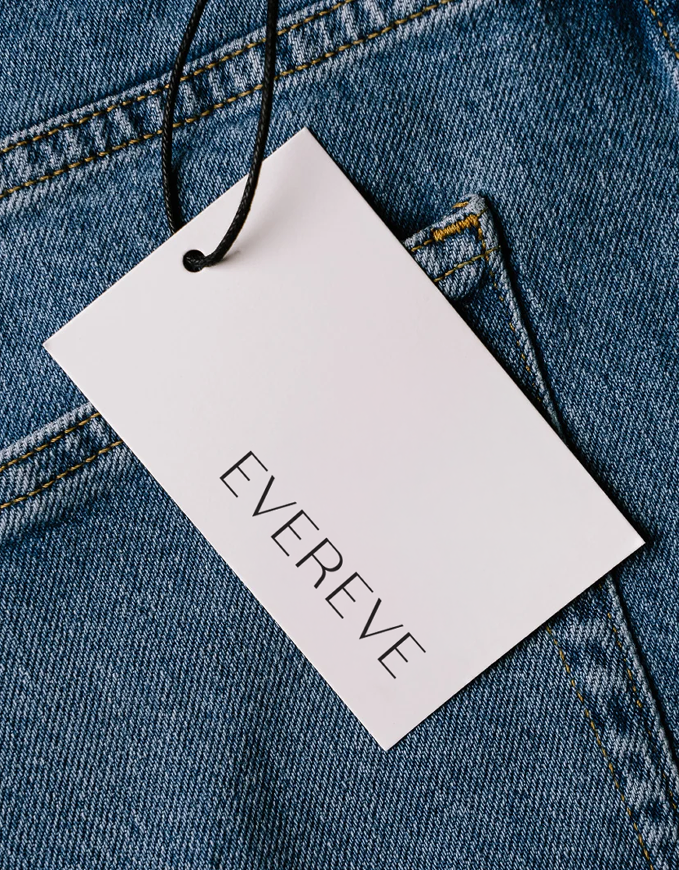 Close-up of a white clothing tag with the partially visible word "EVER" attached to blue denim jeans with orange stitching.