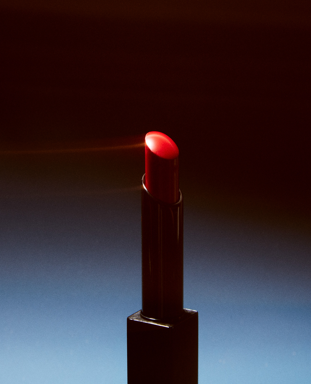 A closed red lipstick in a black case with the cap on, photographed against a dark, gradient background.