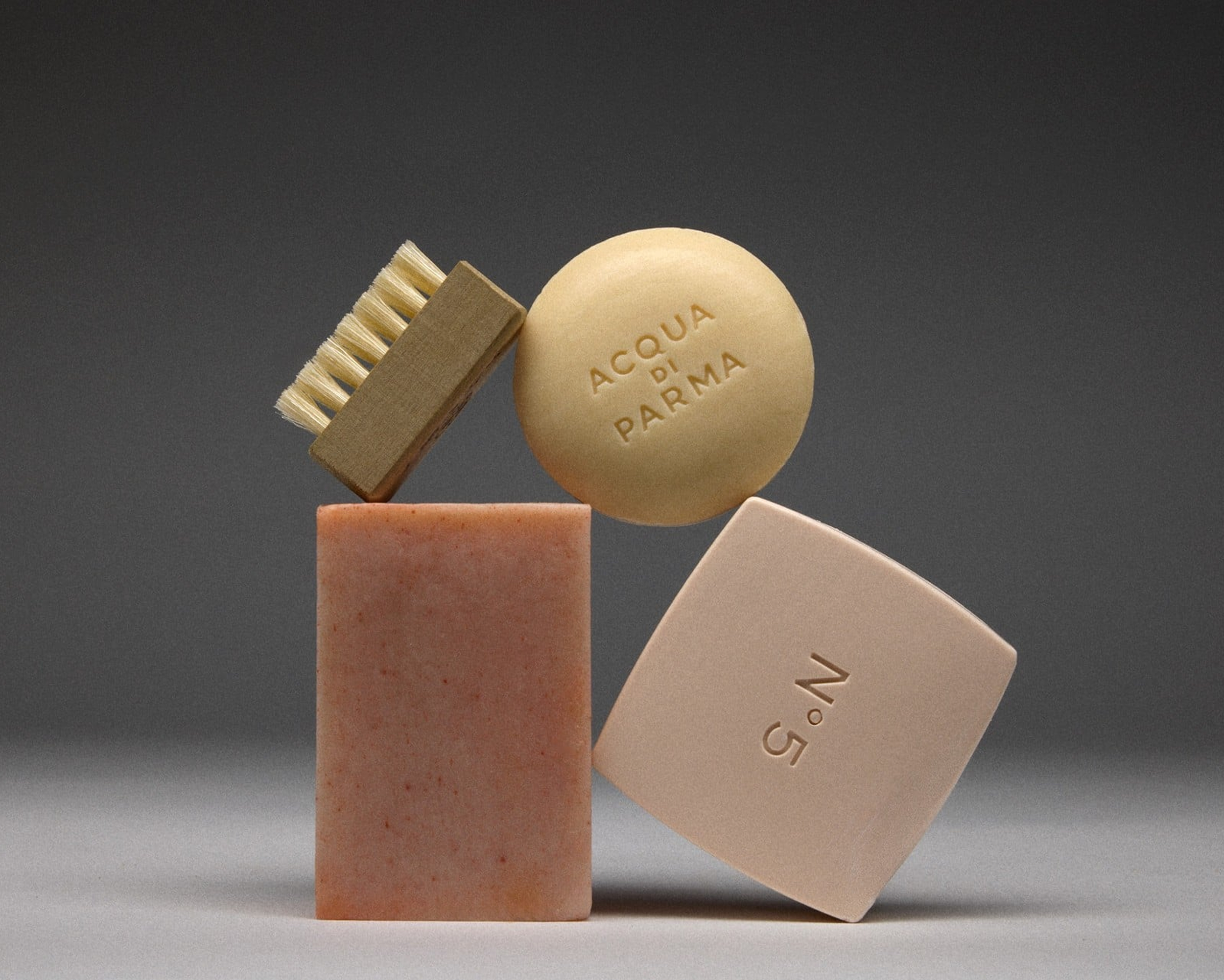 A collage of small objects including a wooden scrub brush, a round beige tablet with the words "ACQUA DI PARMA" engraved, a pink rectangular block, and a white rectangular block with the number "5" engraved, arranged on a flat surface against a dark gray background.