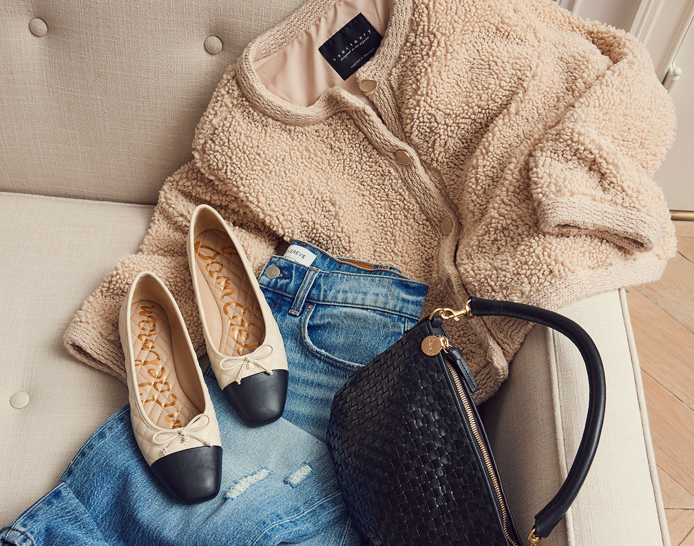 A beige teddy fleece jacket, a pair of beige and black ballet flats, a pair of ripped blue jeans, and a black woven handbag with a gold zipper and strap are arranged on a cream-colored couch.