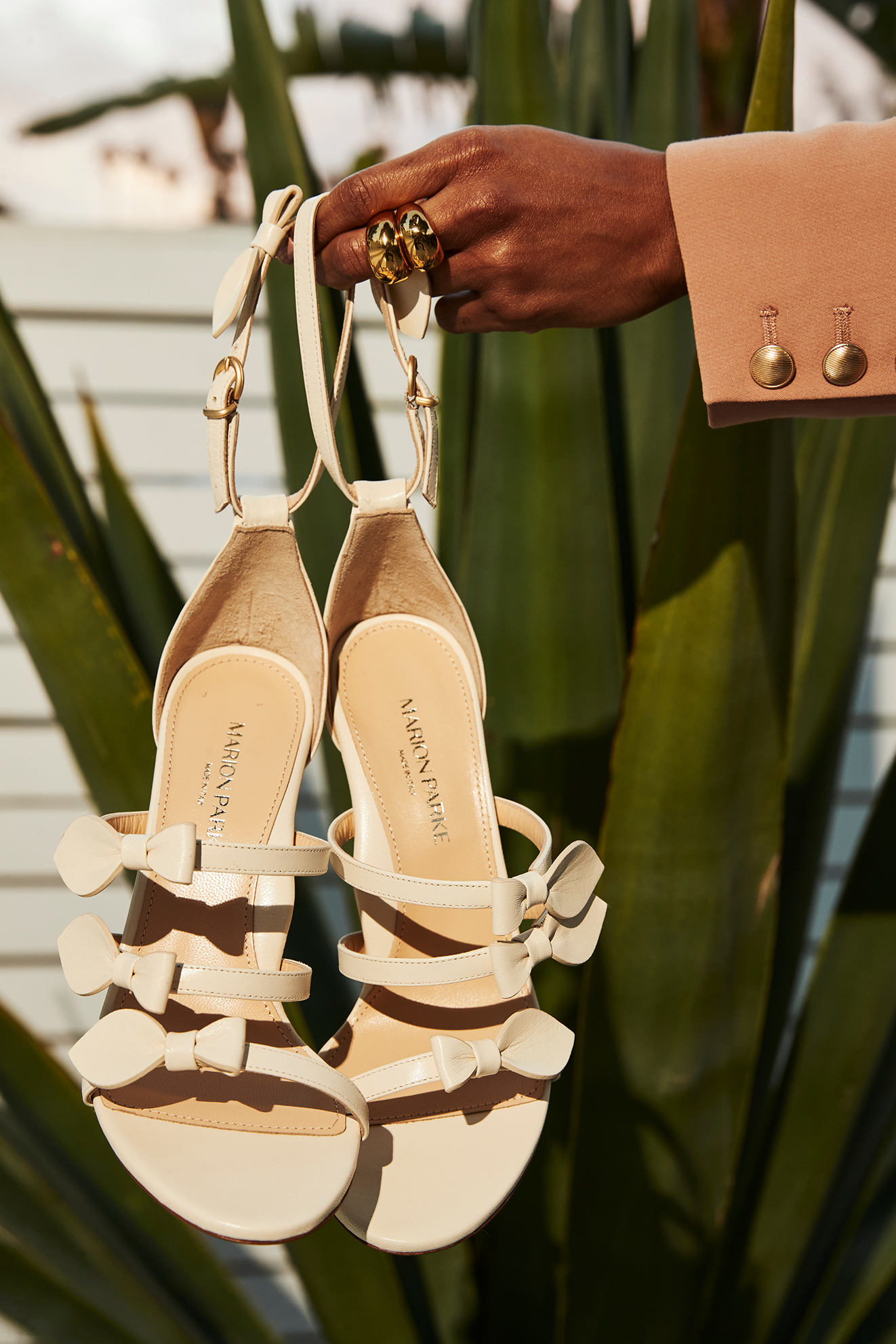 Pair of white high-heeled sandals with bows on the straps hanging on a plant, with a person's hand holding some jewelry in the background.