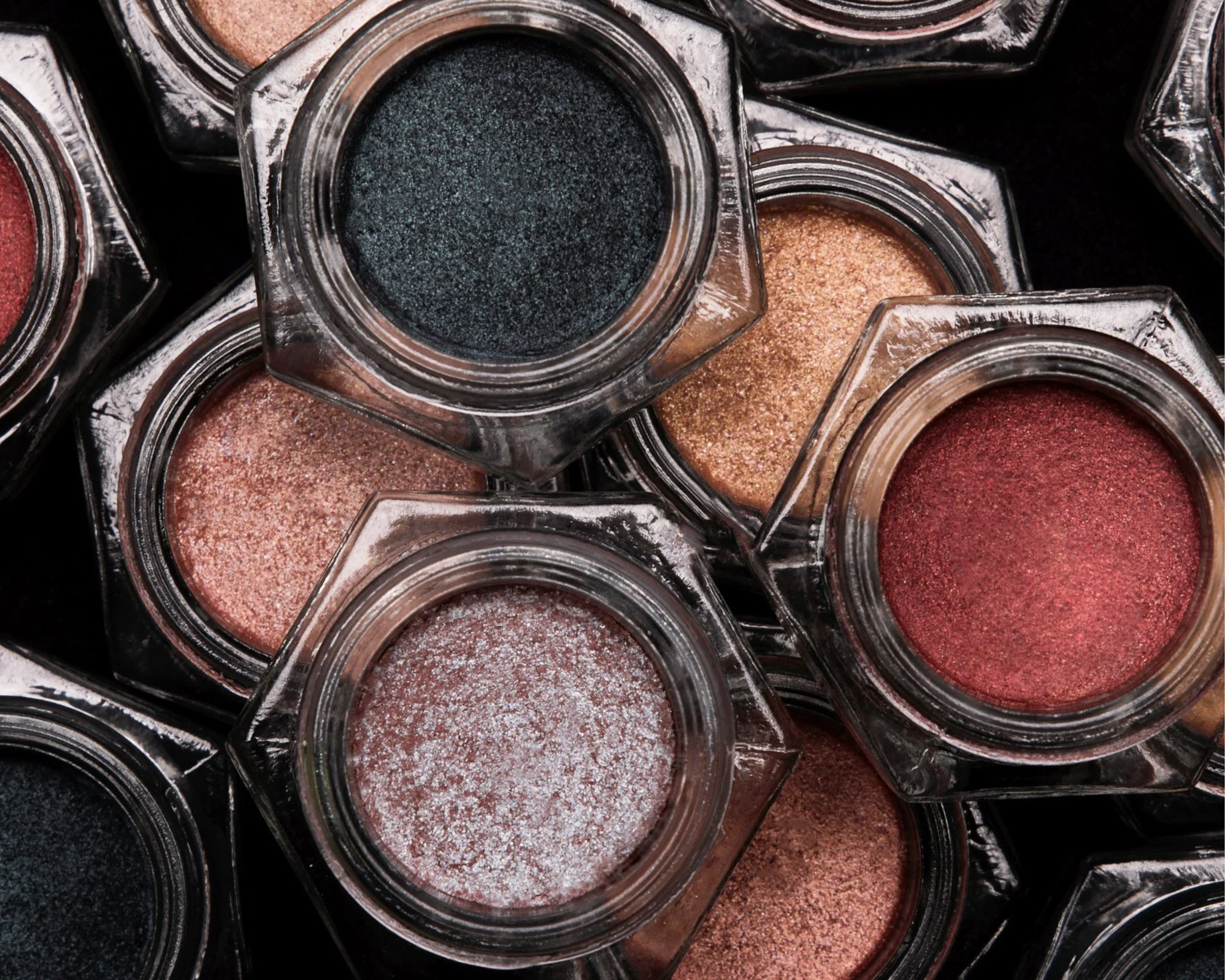 Close-up of multiple open eyeshadow jars with shimmery, textured makeup powders in shades of black, brown, and red.