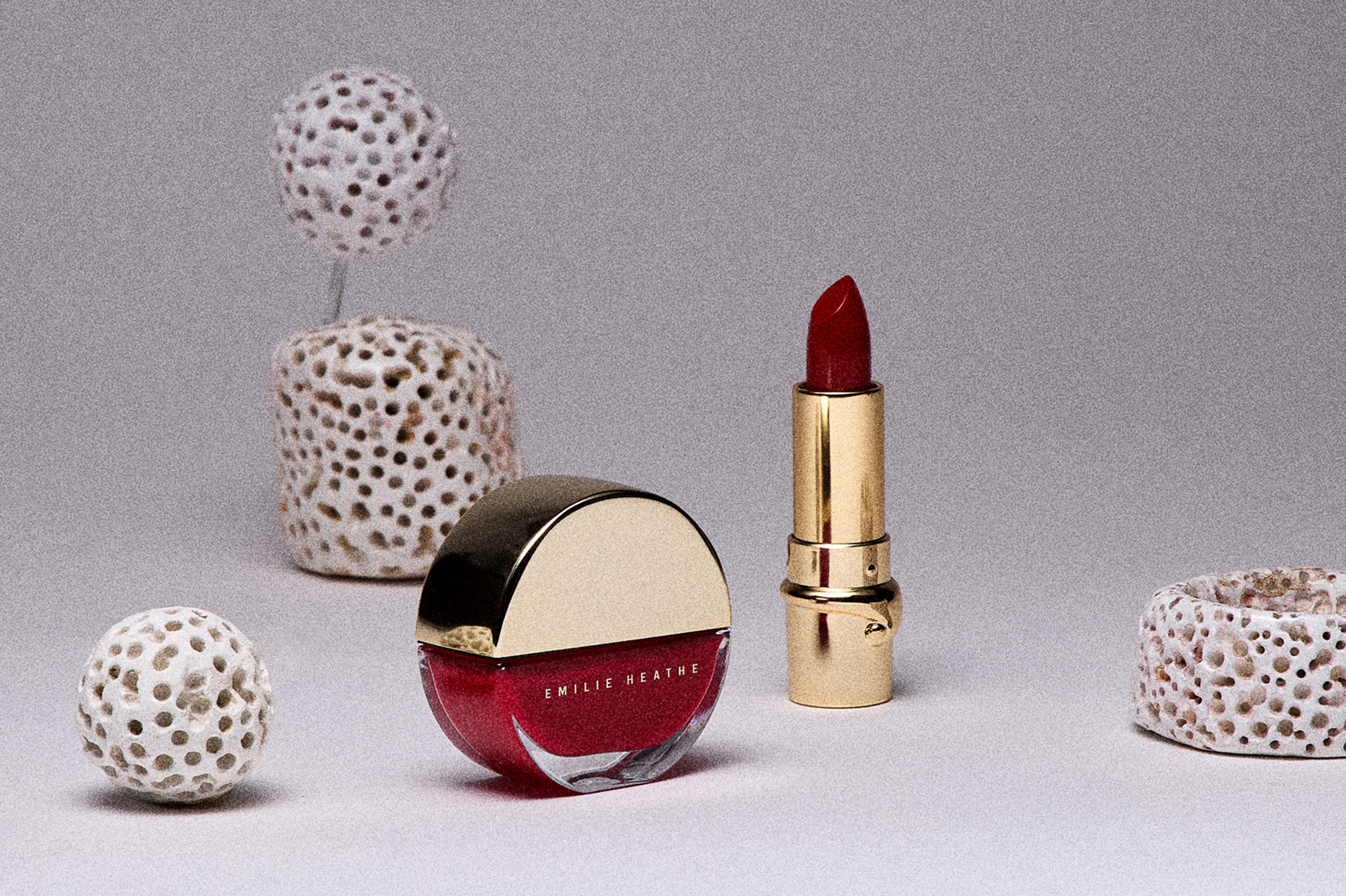Red lipstick in gold tube, dark red nail polish, surrounded by decorative ceramic objects with cut-out patterns.