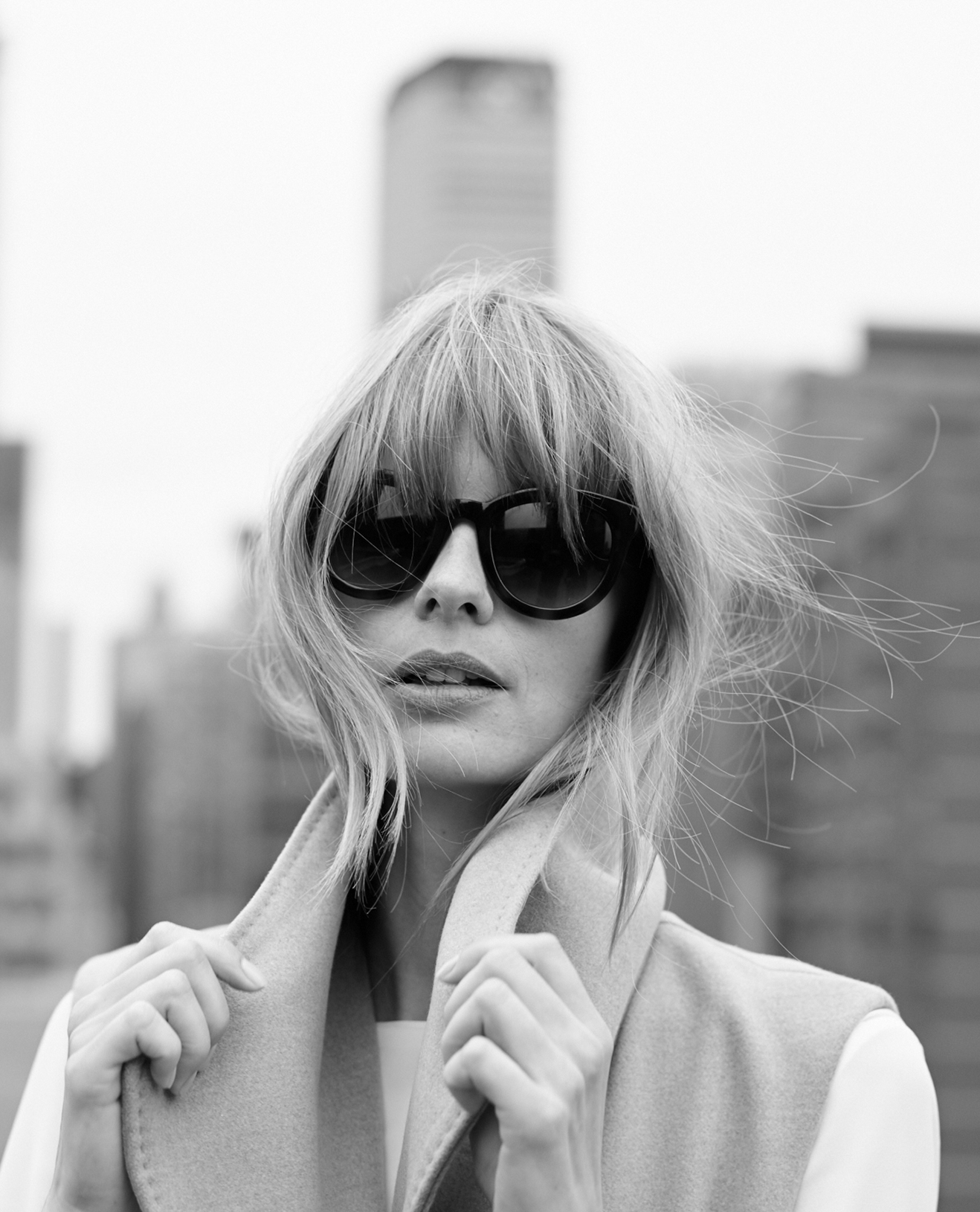 Black and white photo of a woman with blonde hair wearing large dark sunglasses and a light-colored coat, standing outdoors with blurry buildings in the background.