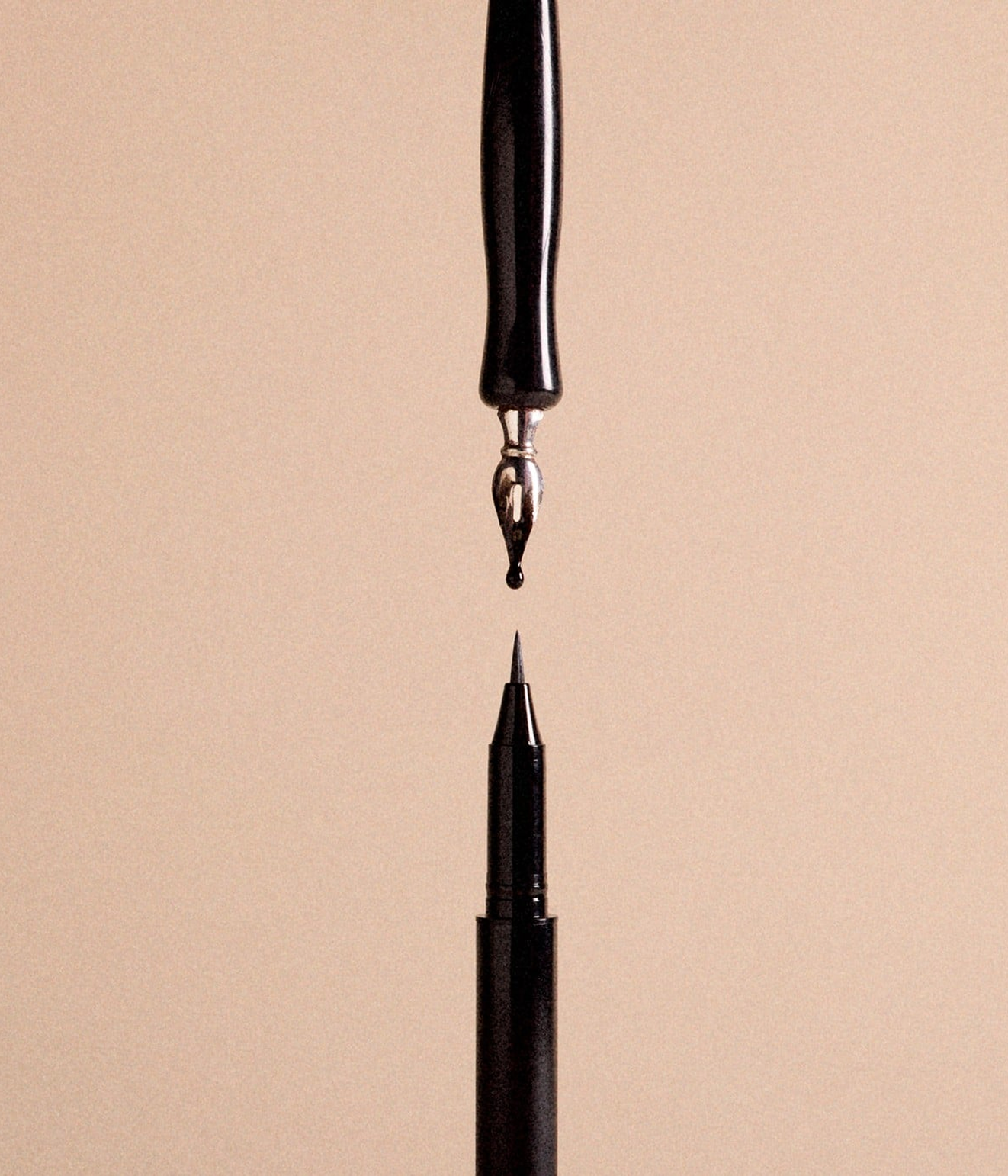 Close-up of two fountain pen nibs facing each other with ink drops forming a reflection on a beige background.