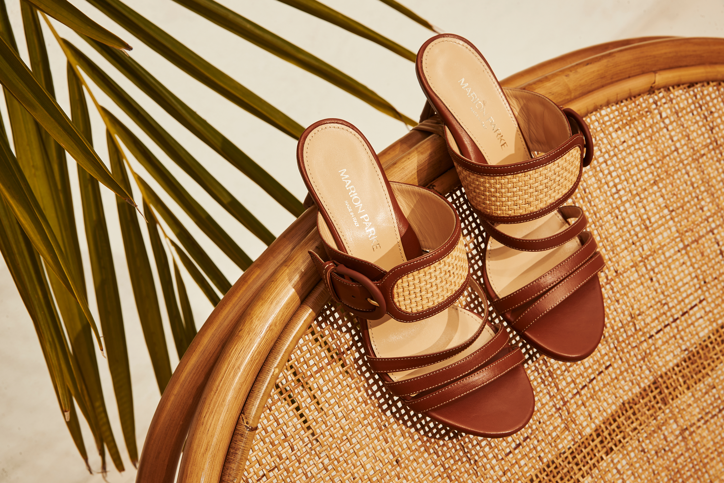 A pair of tan and brown woven leather sandals placed on a circular rattan chair with a woven seat, beside green palm leaves.