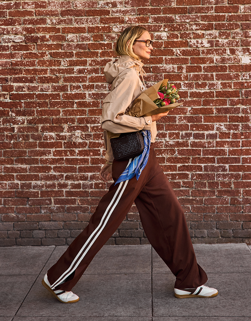 Woman in sunglasses and casual outfit holding a bouquet of flowers, walking past a brick wall.