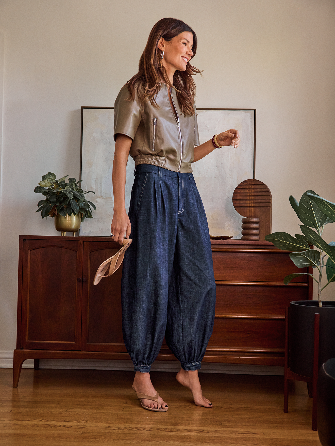 A woman in a tan leather jacket and wide-legged denim pants standing barefoot inside a room with a wooden floor, holding a straw hat, with a mid-century wooden cabinet, potted plants, and abstract artwork in the background.