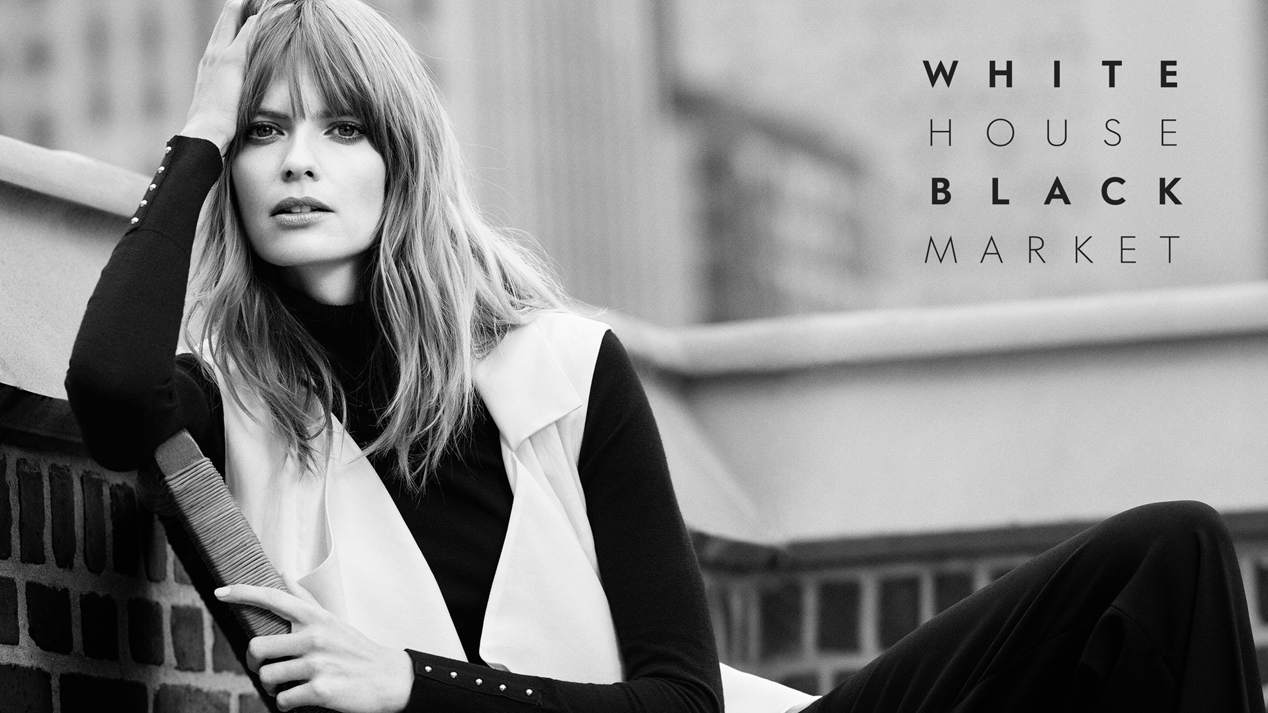 Black and white photo of a woman with long hair sitting against a brick wall, wearing a black and white jacket. The background has an out-of-focus urban setting. Overlaid text reads "WHITE HOUSE BLACK MARKET."