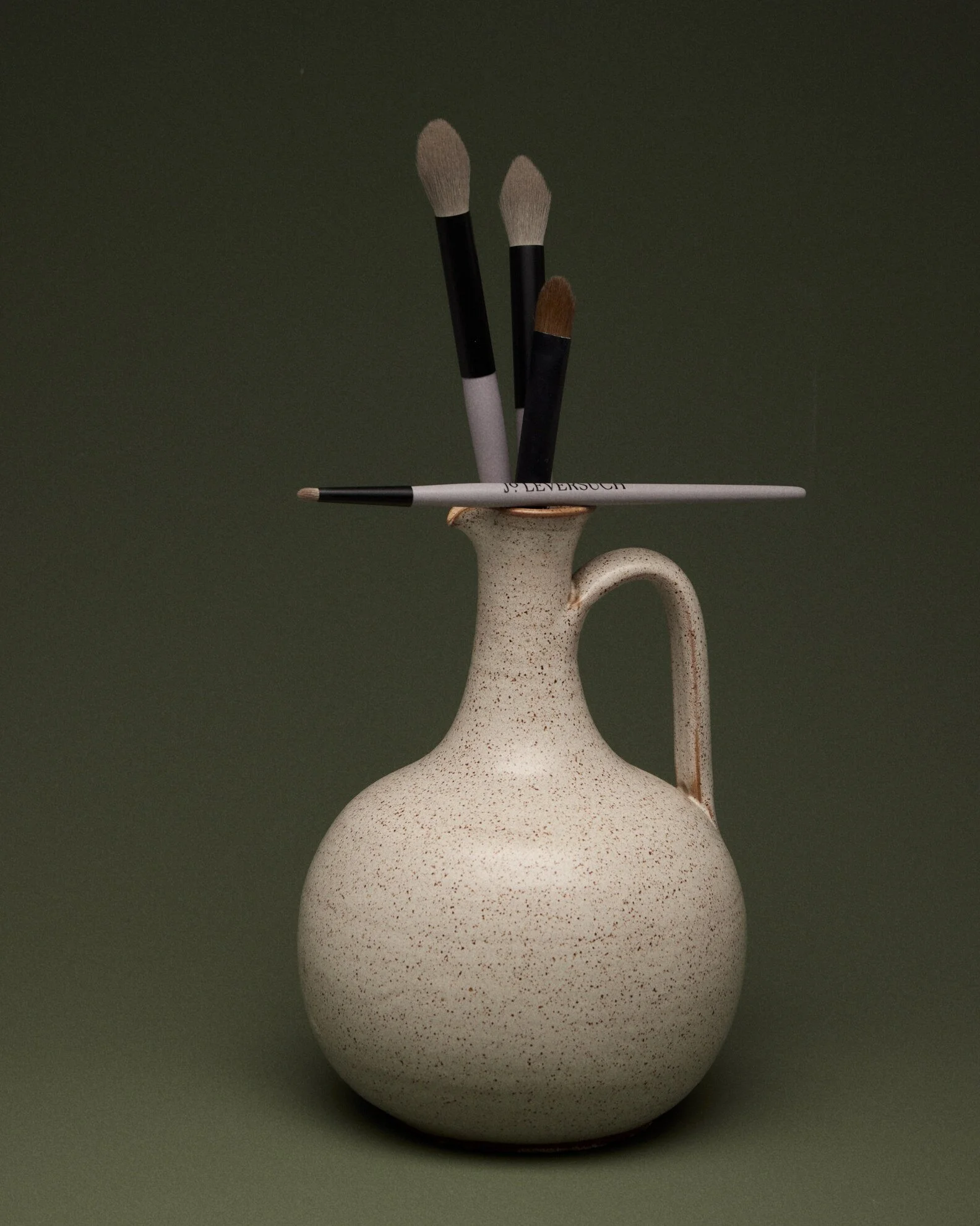A speckled beige ceramic vase with a long neck and an oval-shaped body, holding three makeup brushes with black handles and light-colored bristles against a dark green background.