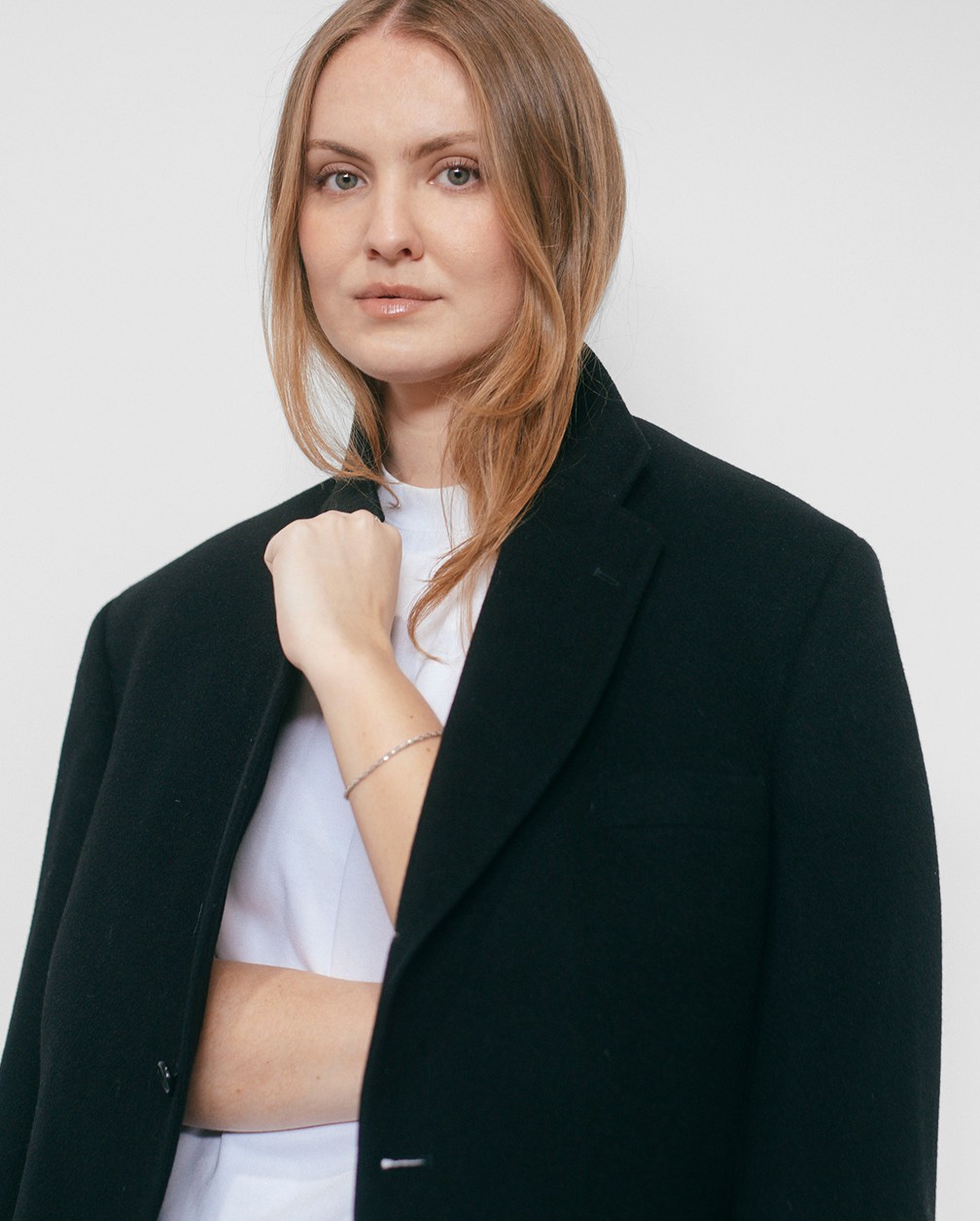A woman with red hair wearing a black blazer, white t-shirt, and a delicate bracelet. She has a serious expression, looking directly at the camera, with a neutral background.