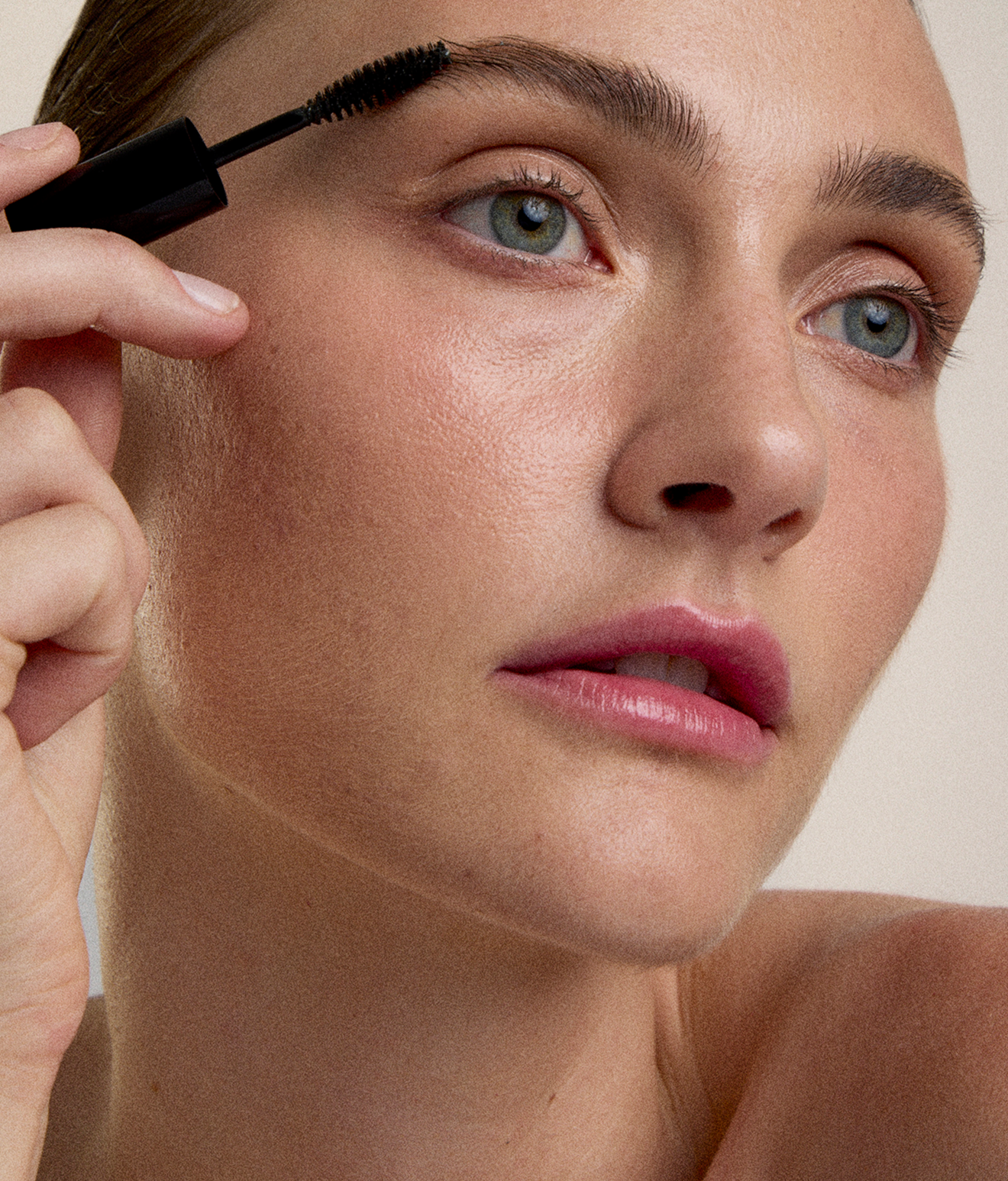 Close-up of a woman applying mascara to her eyelashes, with her face visible and her eyes looking slightly to the side.