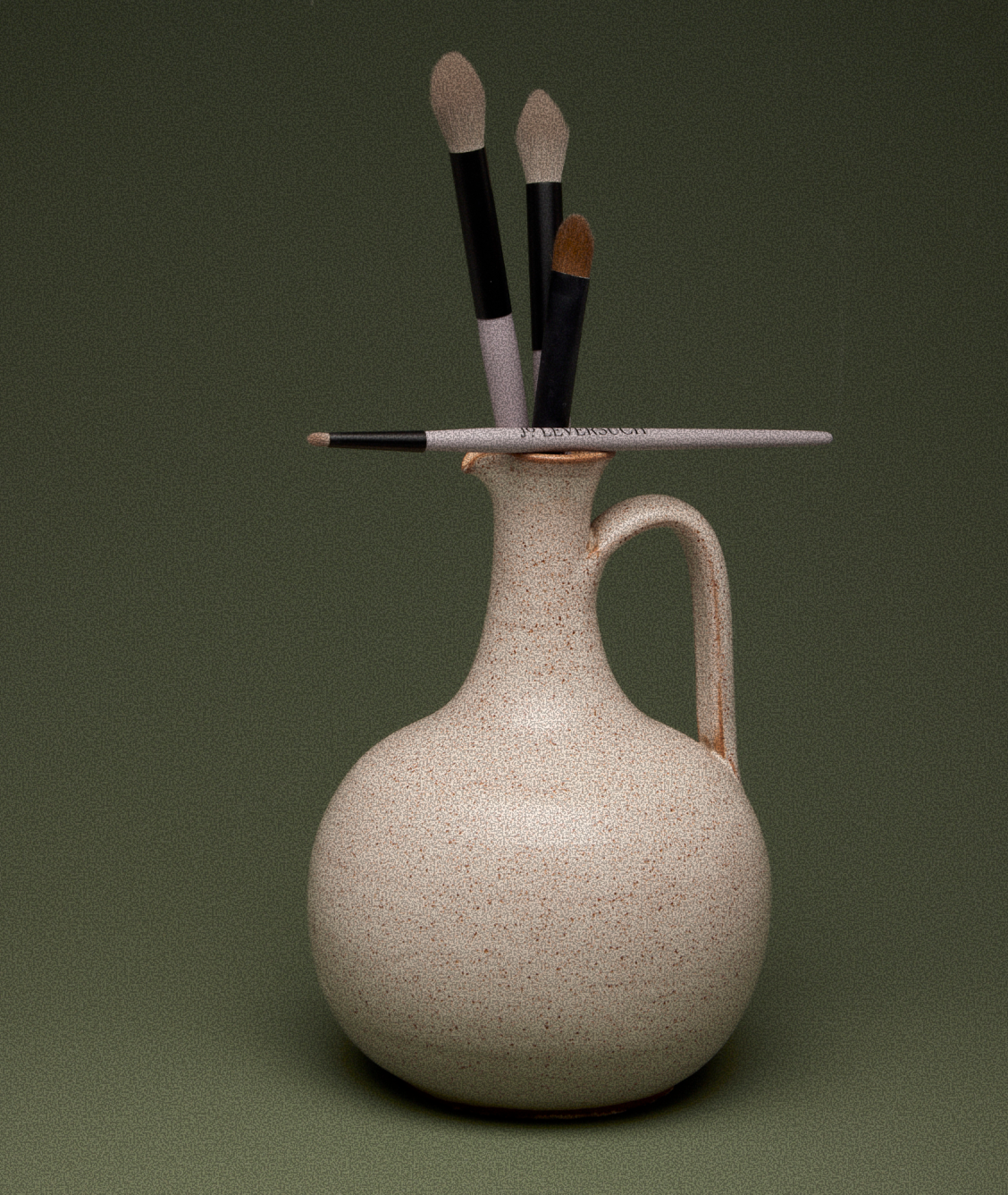 A ceramic jug with a rounded body, narrow neck, and handle, holding three makeup brushes and a eyeshadow brush.