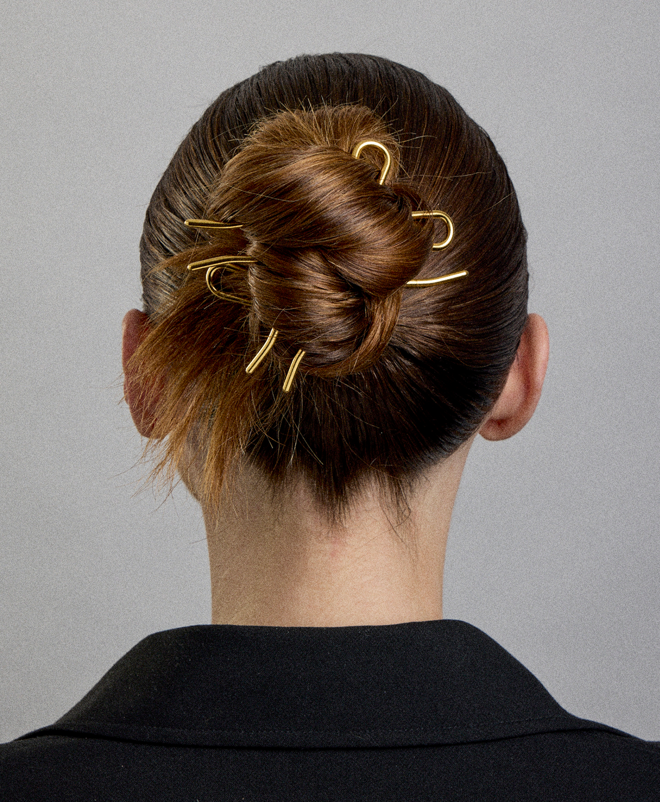 Back of a person's head with brown hair styled in an elegant bun secured with gold hairpins, wearing a black top.